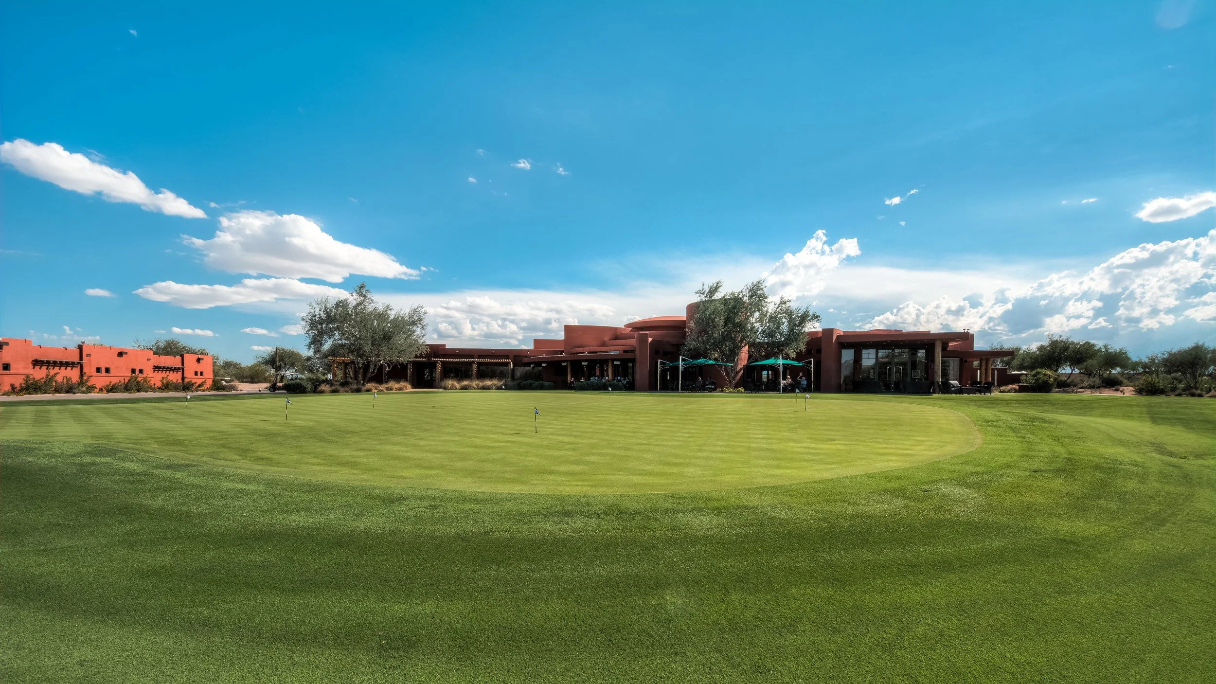 A golf course with a green in front and a modern building in the background, under a blue sky with scattered clouds.