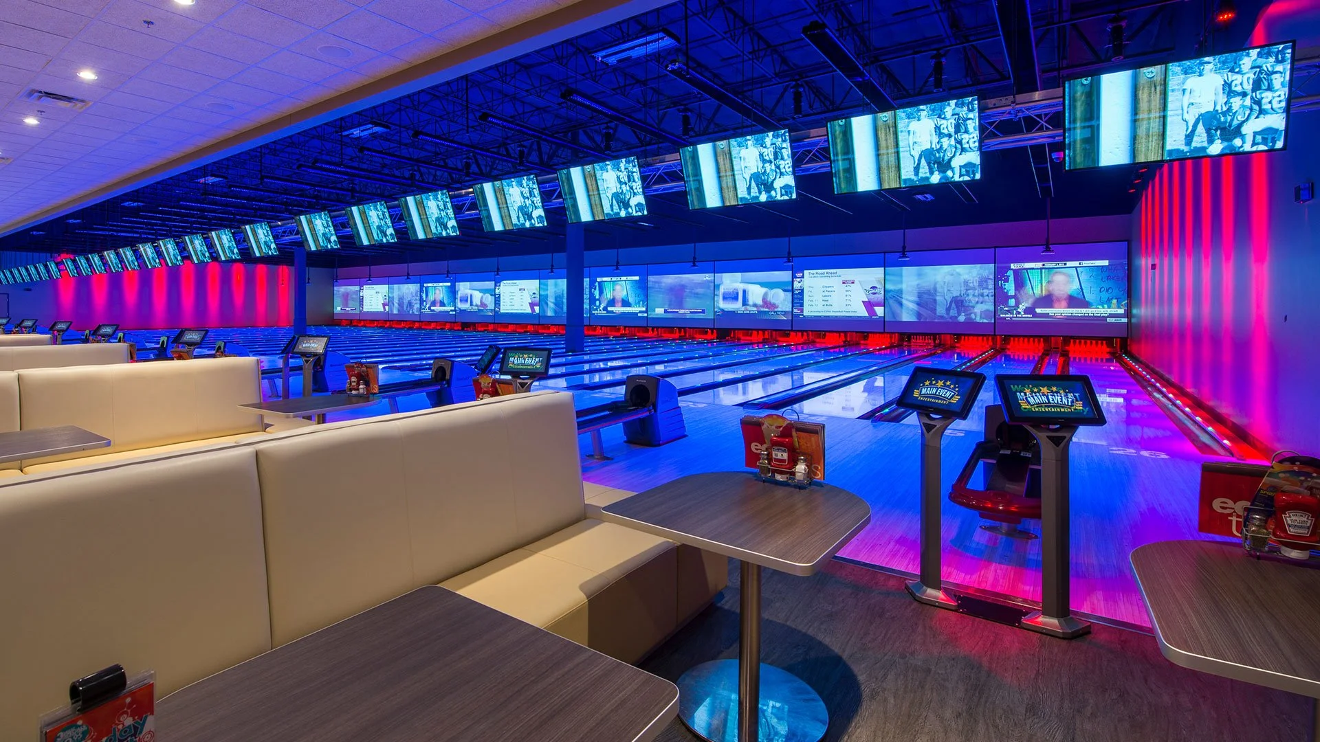 Empty bowling alley with multiple lanes, large screens above, seating booths in the foreground, and a neon-lit wall in the background.