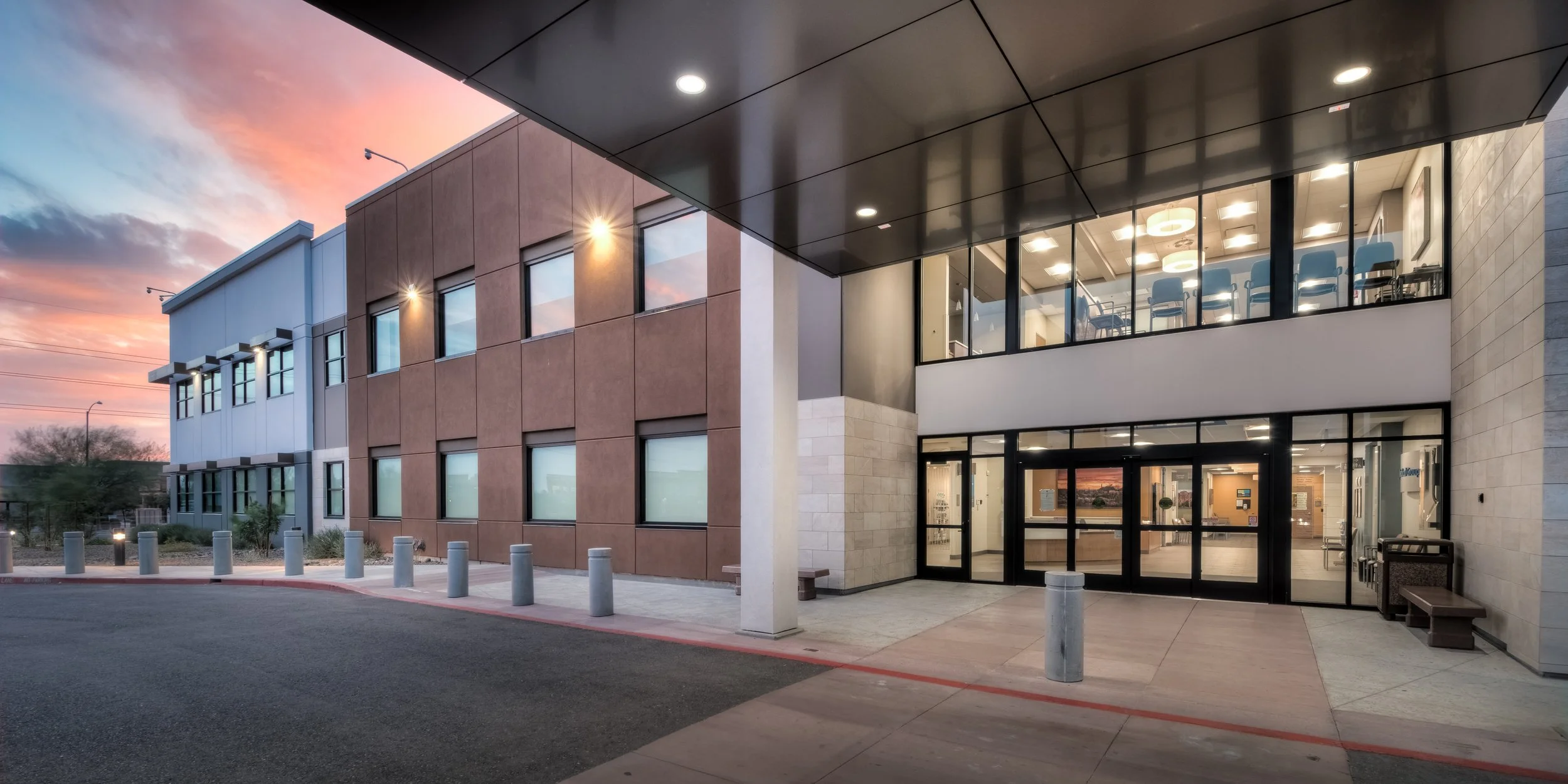 Exterior of a modern multi-story building at sunset, with large glass entrance doors and windows, outdoor lighting, and a sidewalk with safety bollards.