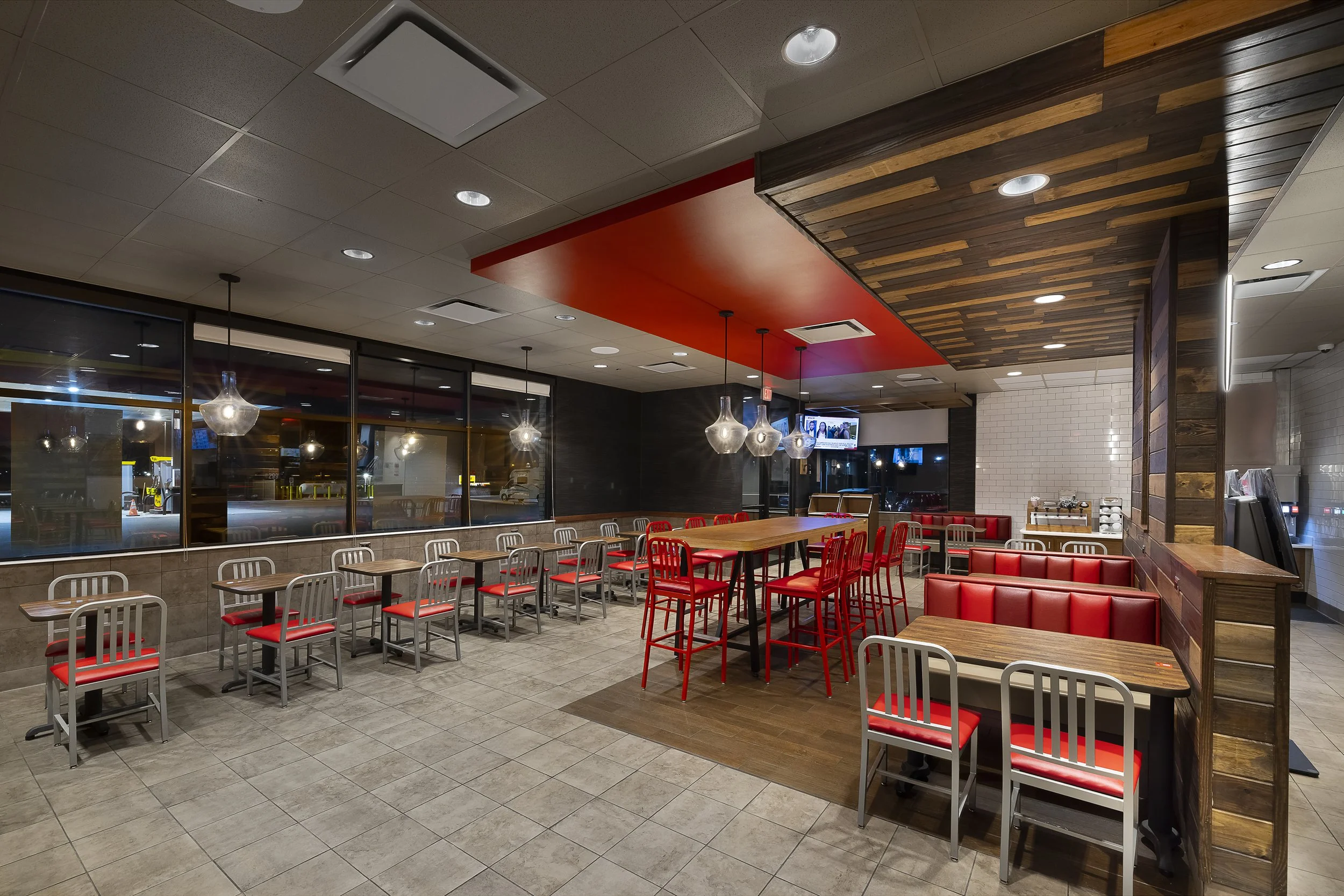 Empty restaurant with tables, chairs, and booths, illuminated by hanging pendant lights, with a large window, TV screens on the wall, and a coffee station in the background.