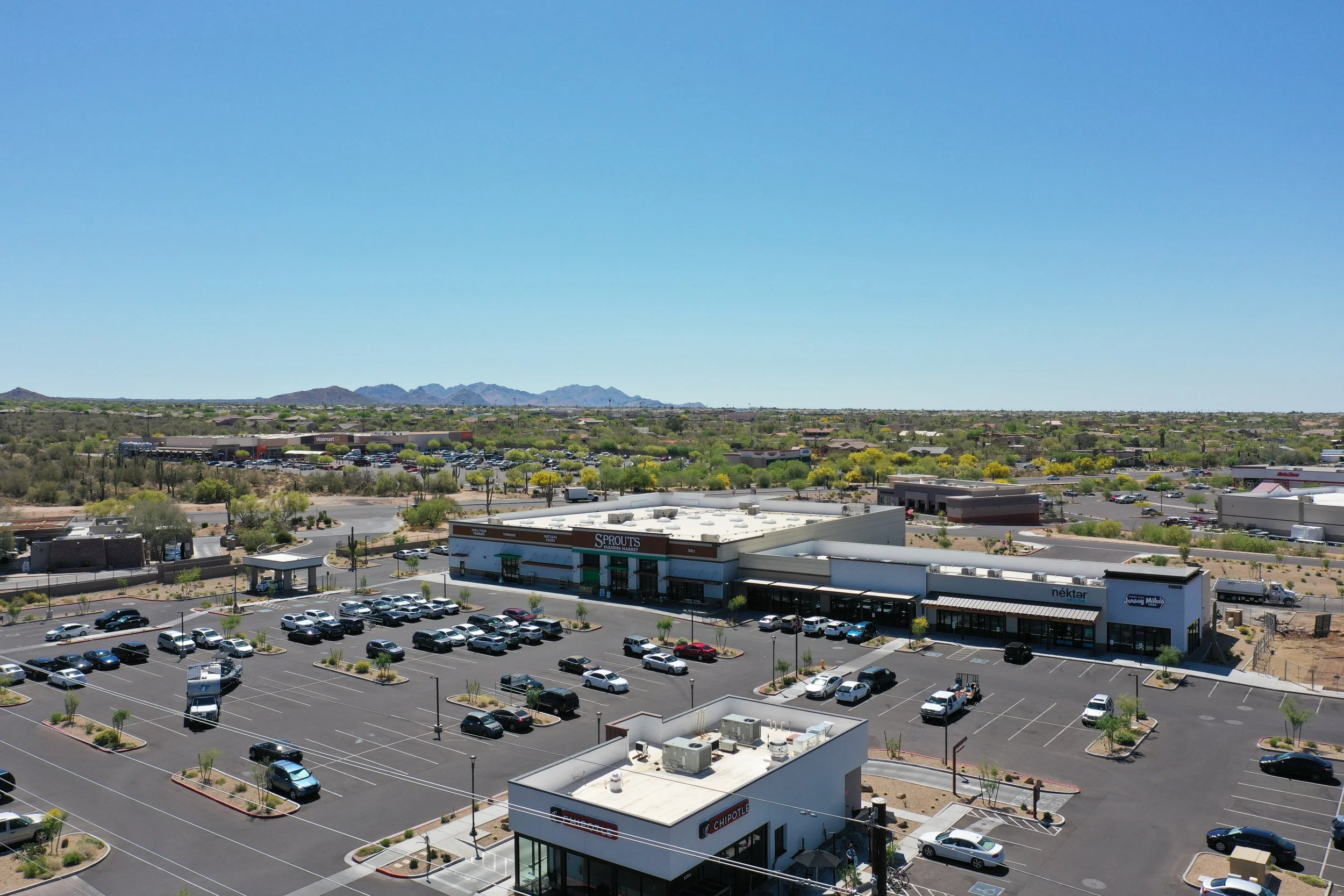 A wide view of a retail parking lot with numerous cars, surrounding shops including Chipotle, Sprouts Farmers Market, and Nekter, under a clear blue sky with distant mountains on the horizon.