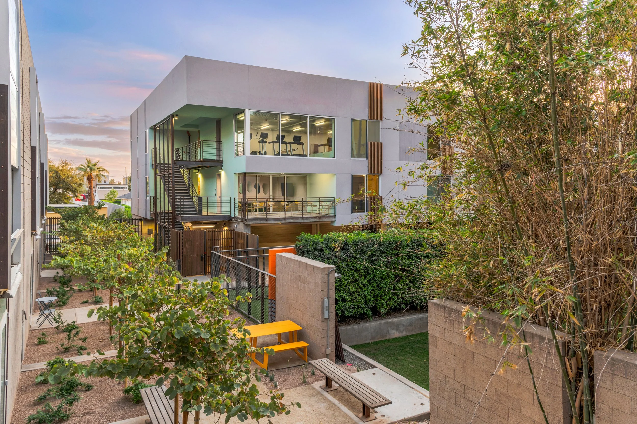 Modern multi-story apartment building with large windows, outdoor balconies, and stairs, surrounded by landscaped outdoor area with trees, benches, and a yellow picnic table, during sunset.