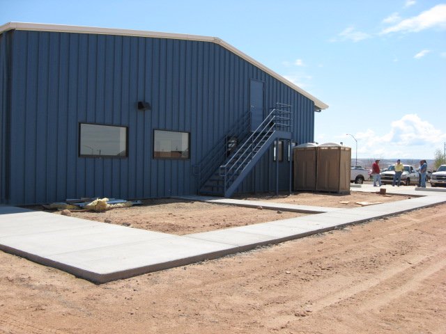 A blue metal building with a stairway, located on a construction site with a concrete sidewalk and portable toilets, surrounded by parked cars and a clear blue sky.