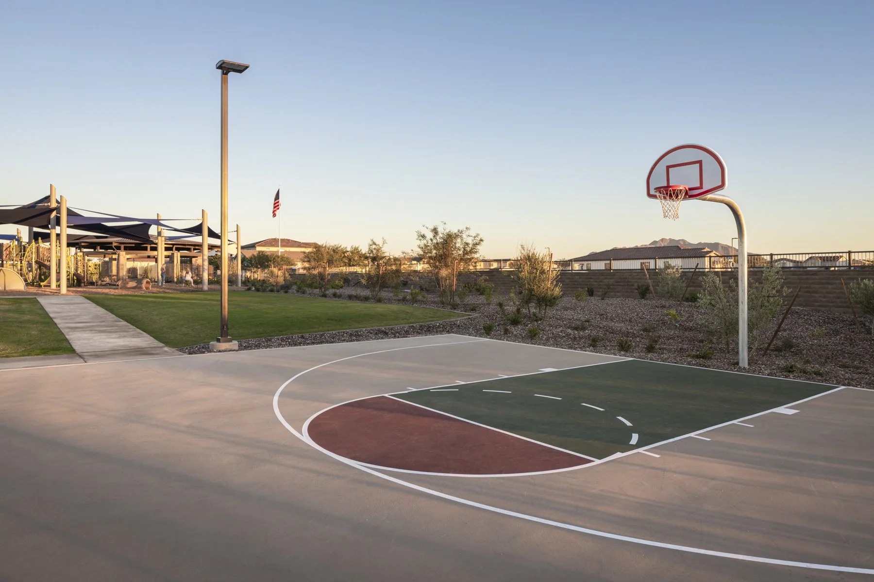 An outdoor basketball court with a hoop and backboard, surrounded by a landscaped area with rocks and small bushes, in a suburban neighborhood during sunset.
