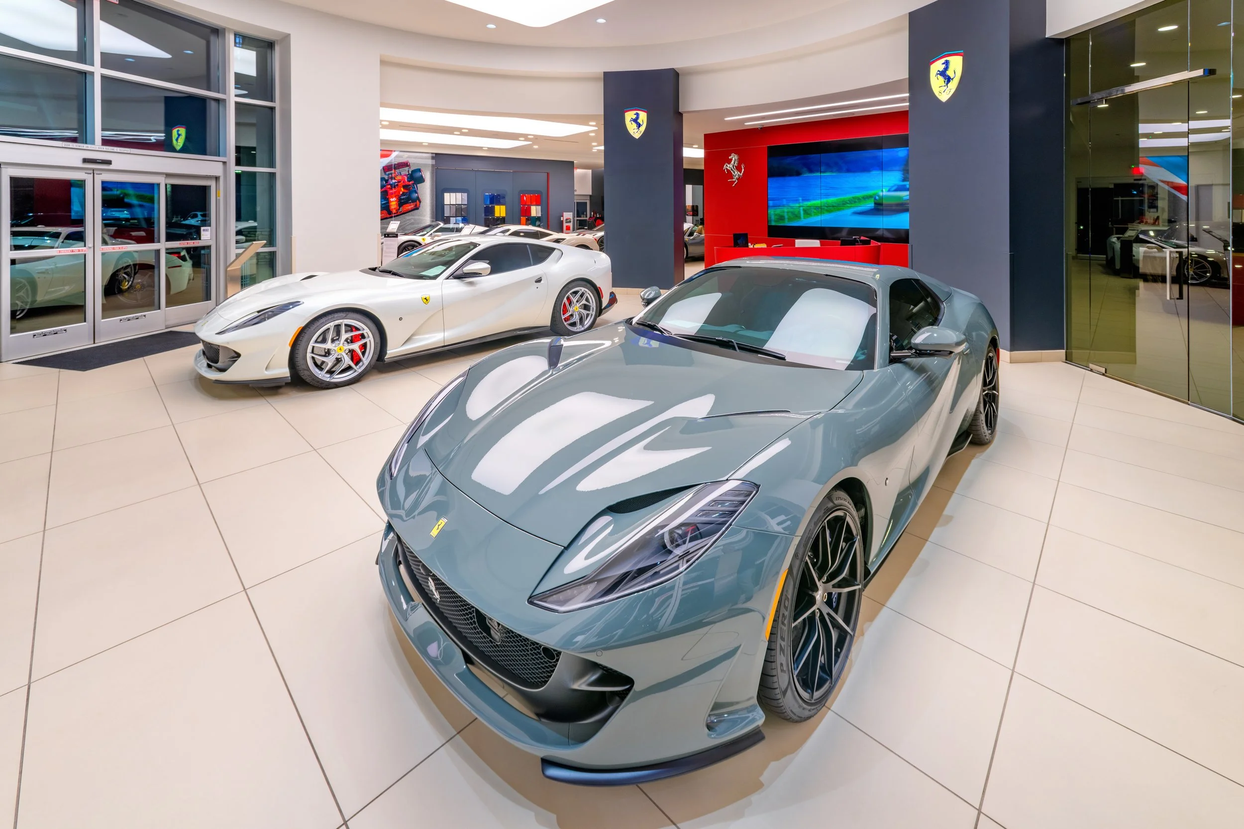 Interior of a Ferrari car dealership showcasing two sports cars, a gray Ferrari and a silver Ferrari, with Ferrari logos on the walls and a digital display in the background.