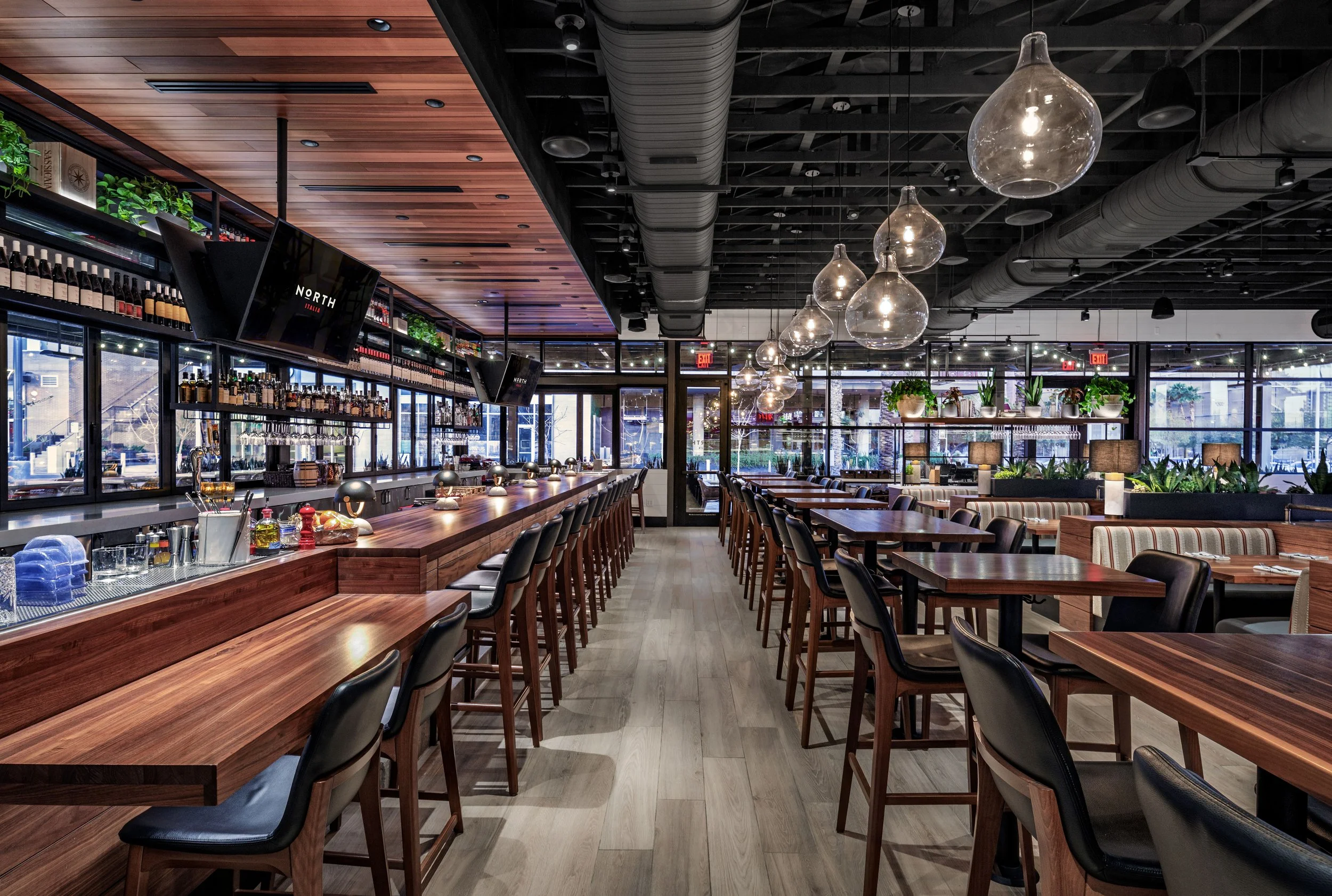 Empty modern restaurant with a bar on the left, wooden tables and black chairs, glass pendant lights, large windows, and potted plants.