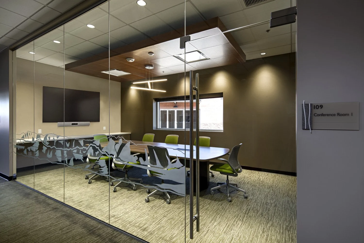 Conference room with glass walls, a large table surrounded by green chairs, a wall-mounted TV, and modern ceiling lighting.