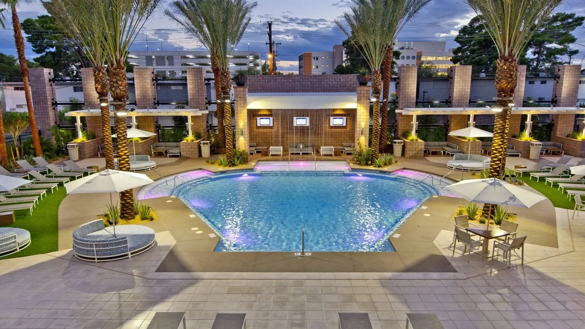 A rooftop swimming pool area at dusk with lounge chairs, umbrellas, palm trees, and illuminated water features.