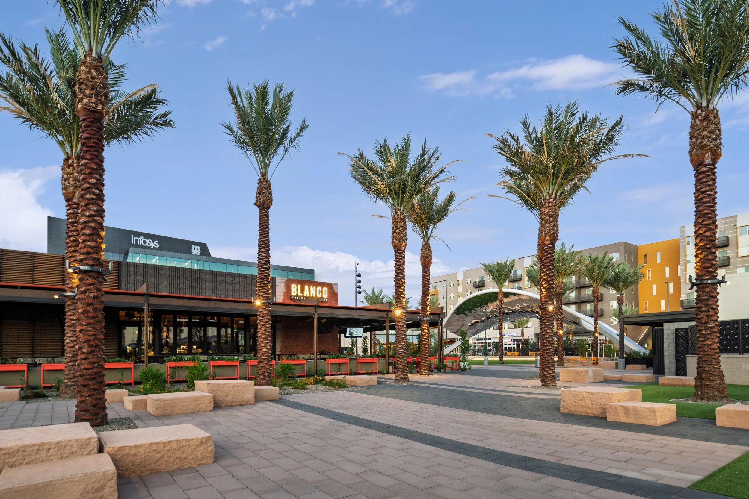 An outdoor shopping area with tall palm trees lining a paved walkway, modern buildings in the background, including a restaurant called BLANCO, and a blue sky with some clouds.
