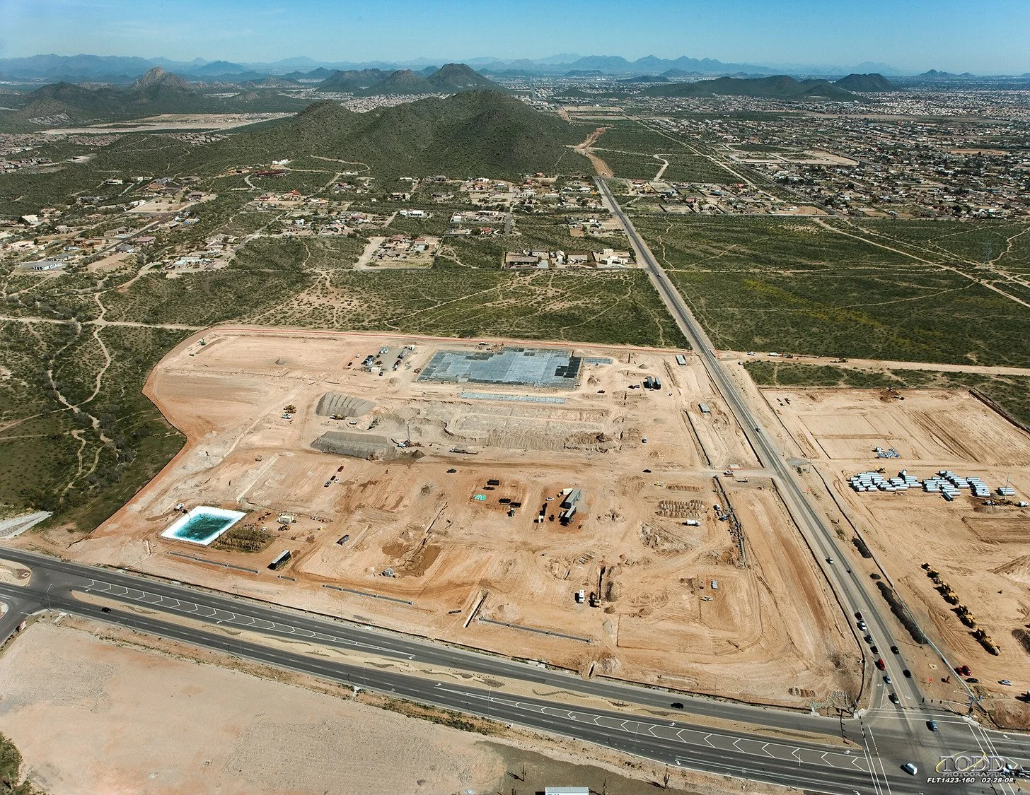 Aerial view of a construction site with dirt and some structures, adjacent to a highway, with a mountain range and residential area in the background.