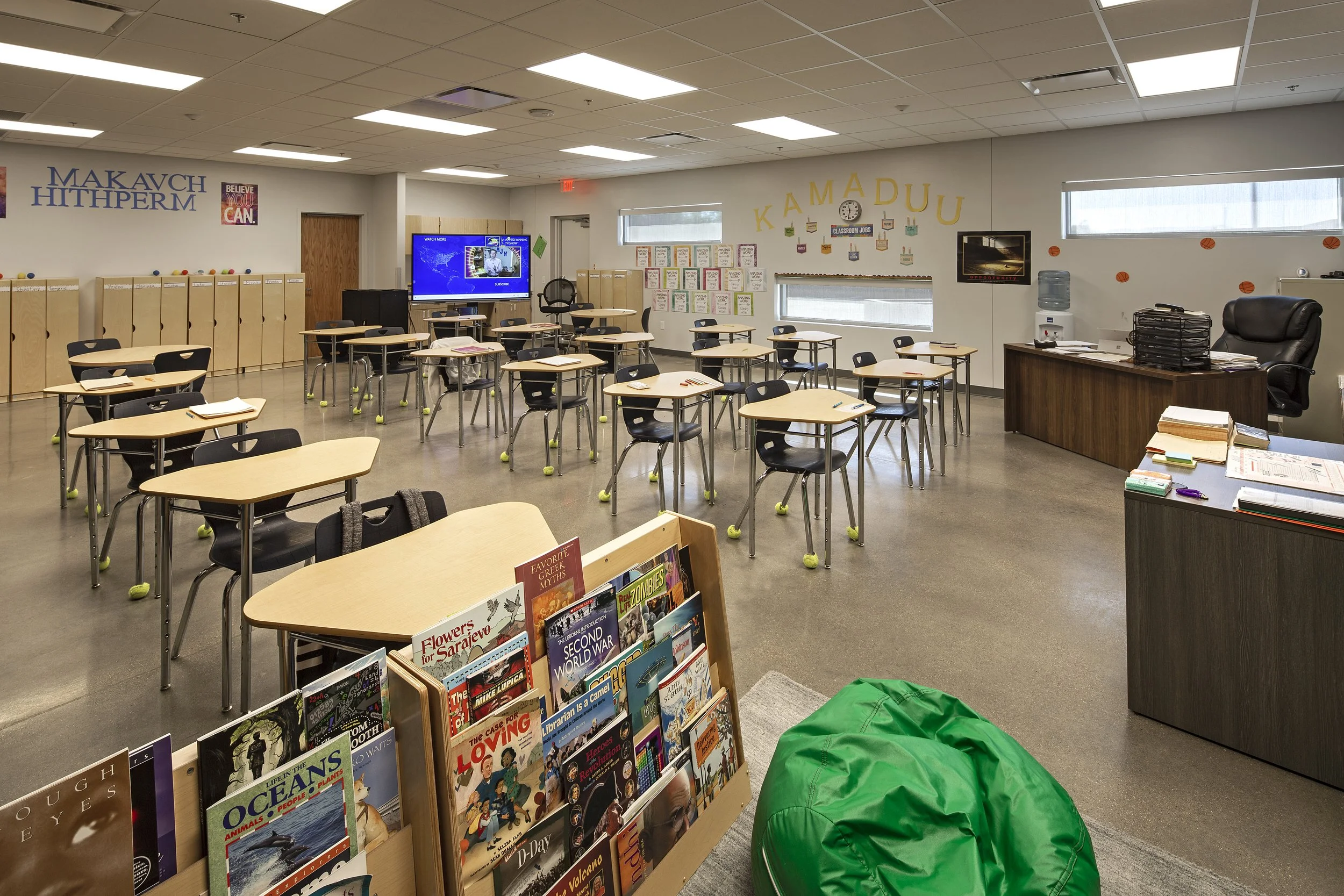 A classroom with desks and chairs, a bookshelf with magazines, a teacher's desk, and bulletin boards on the wall.