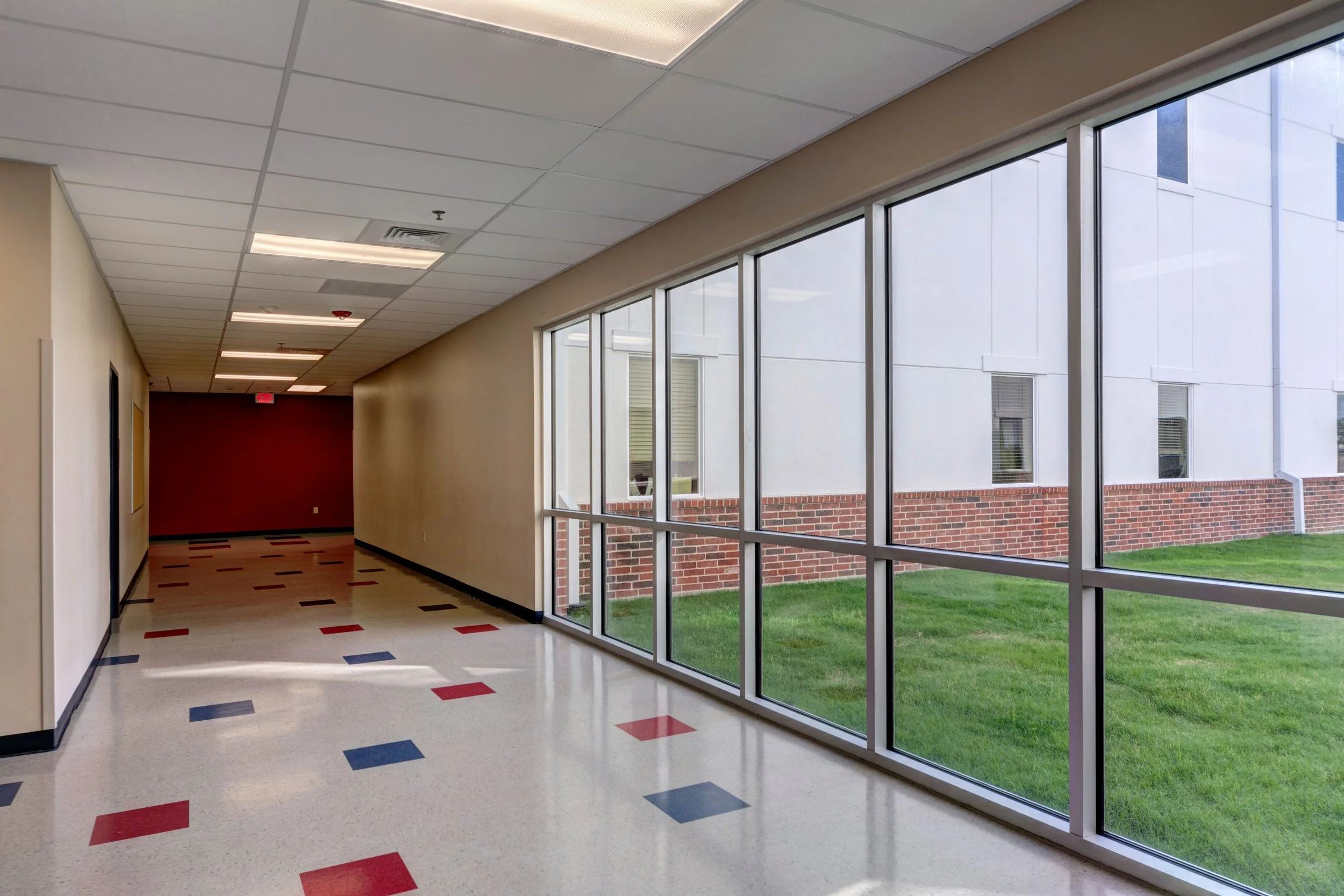 Empty hallway in a building with large windows showing outside grass, with a decorated floor and ceiling lights.