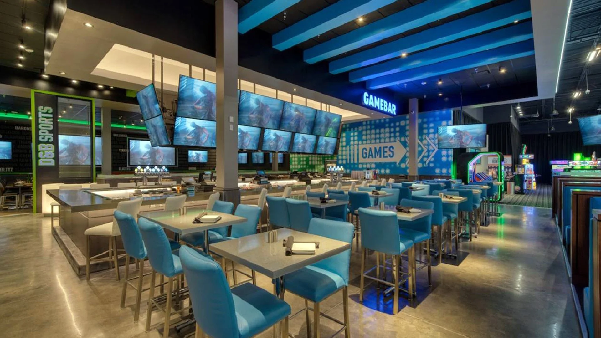 Inside a modern gaming restaurant with multiple large screens displaying video game images, blue cushioned chairs, tables set with napkins and utensils, and arcade game machines in the background.