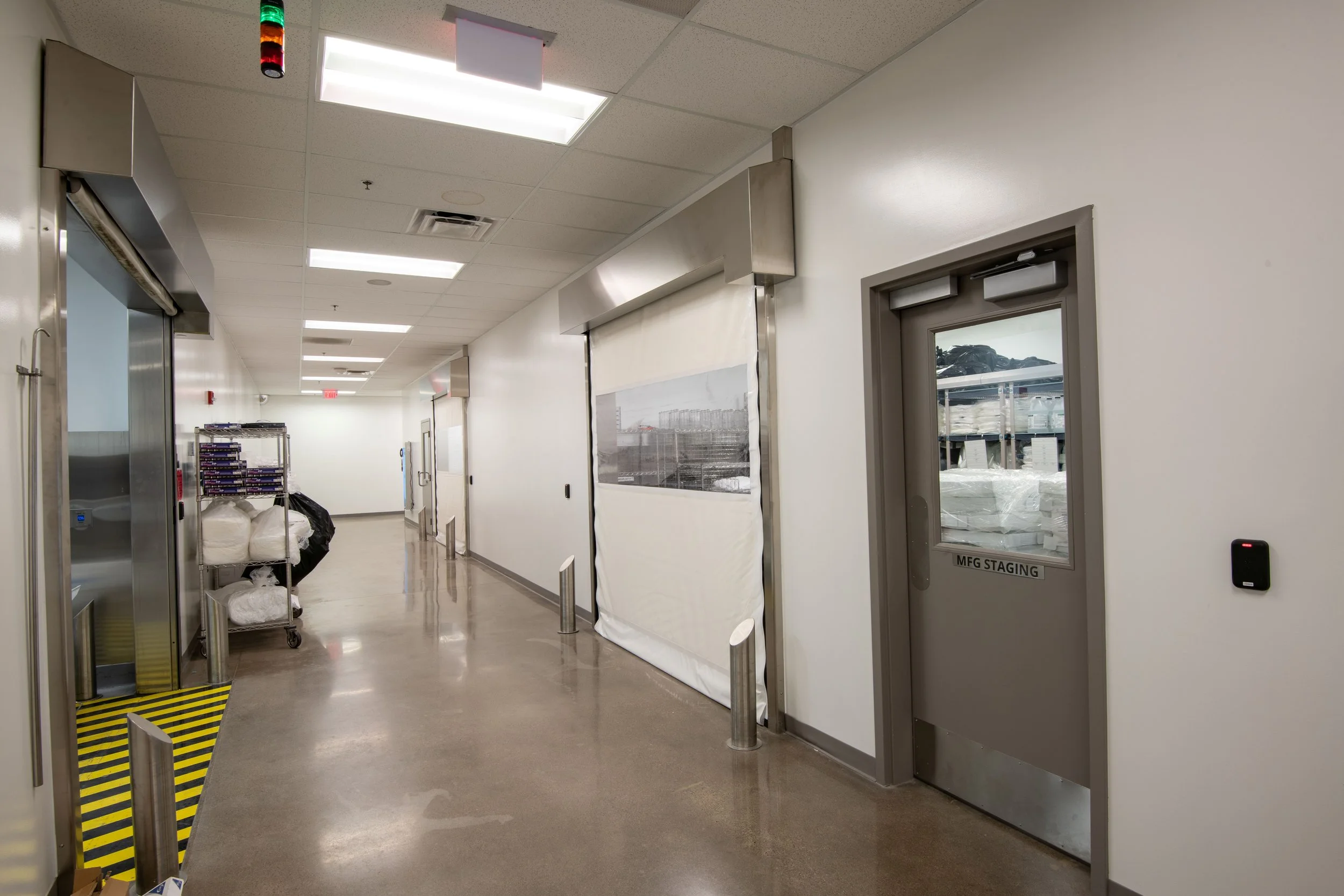 An industrial or commercial building corridor with white walls and a brown polished floor. There is an elevator on the left, a rolling cart with packaged items, and a door labeled 'MFG STAGING' on the right. Fluorescent ceiling lights illuminate the 
