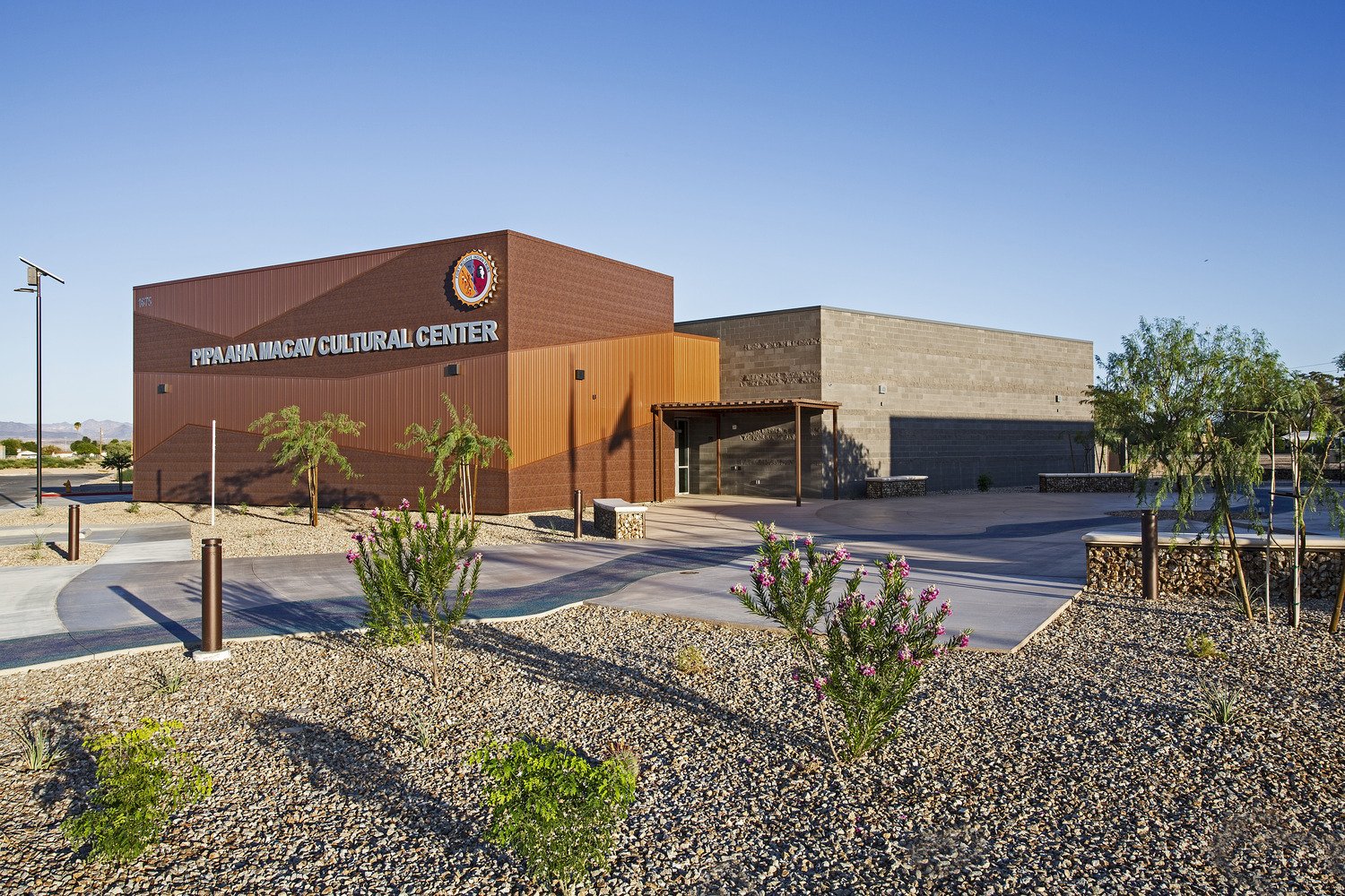 Exterior view of Pipaaha Macav Cultural Center with desert landscaping, small trees, and pink flowering plants in front, under a clear blue sky.