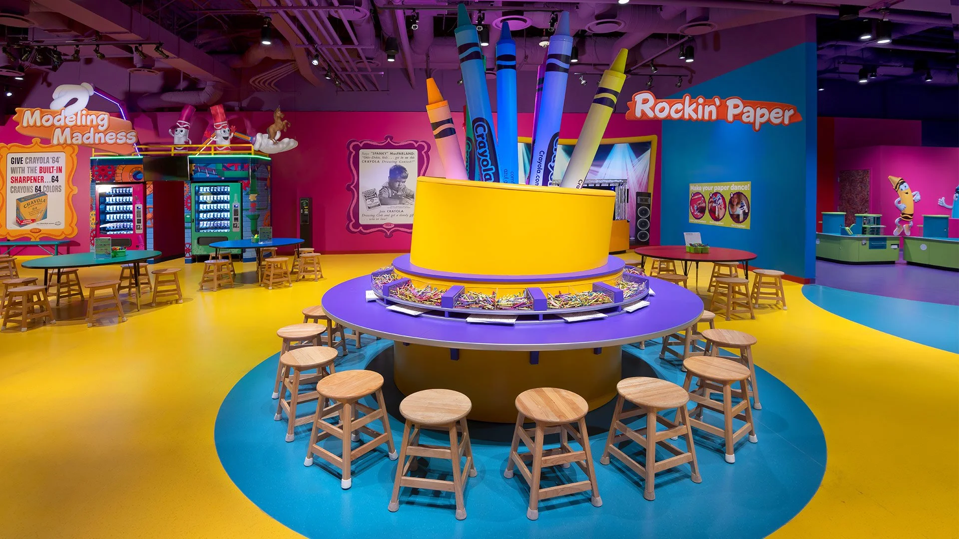 Colorful indoor play area with tables and stools, featuring a large yellow and purple circular display with Crayola crayons and supplies, and vibrant walls with signs and characters, including a pink wall with modeling and paper crafting themes.