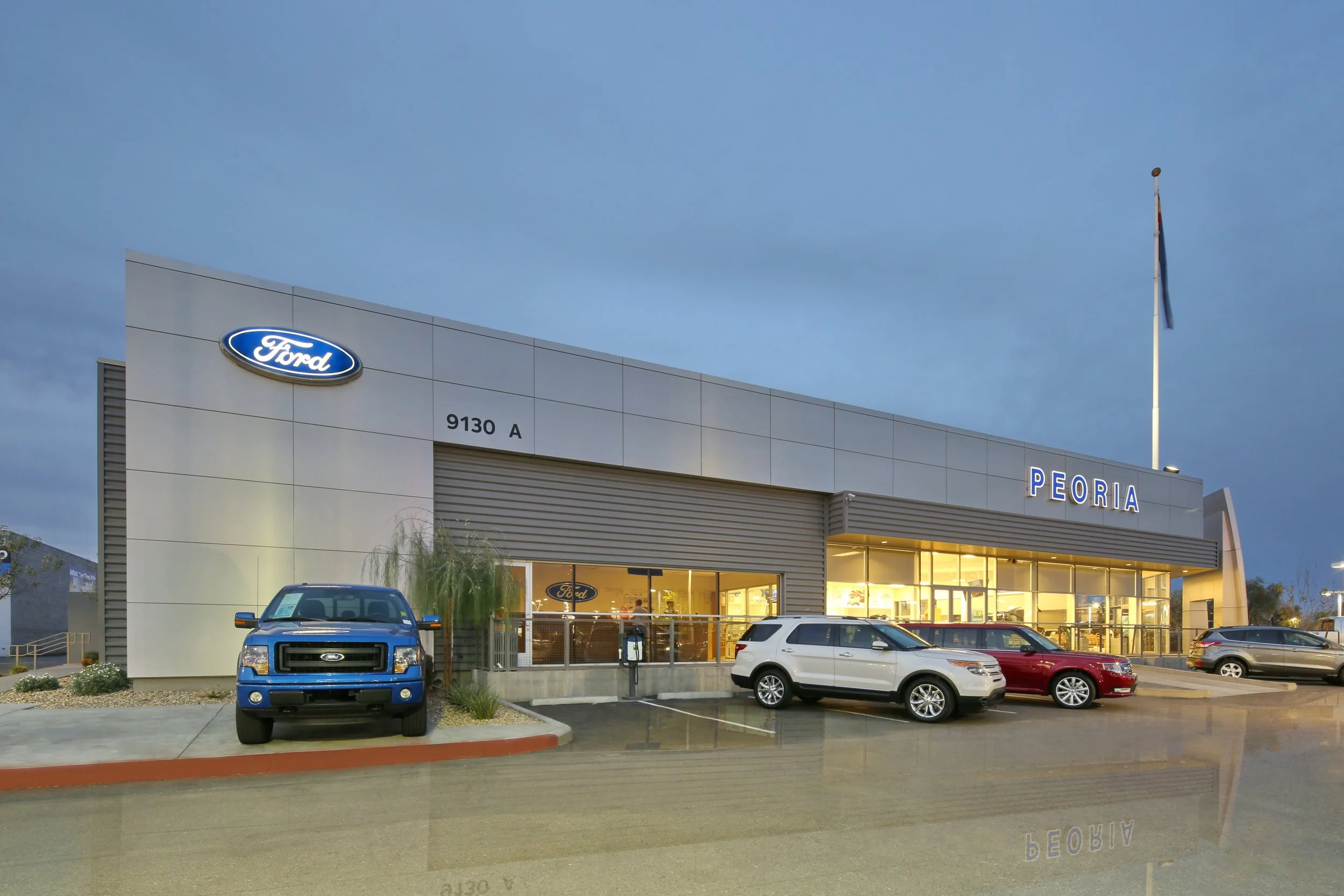 Exterior of a Ford dealership with the Ford logo, the word 'PEORIA', several parked cars, and a flagpole with a flag, during dusk or evening.