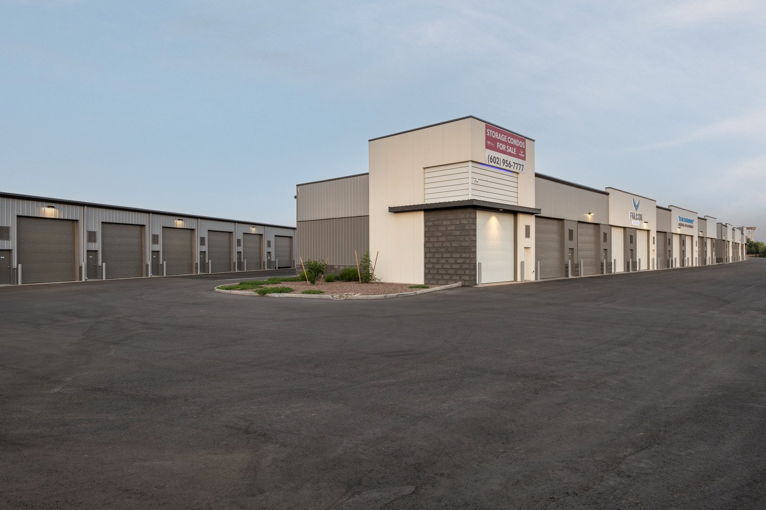 A large storage facility with multiple units, a sign indicating storage condos for sale, and a wide asphalt driveway under a cloudy sky.