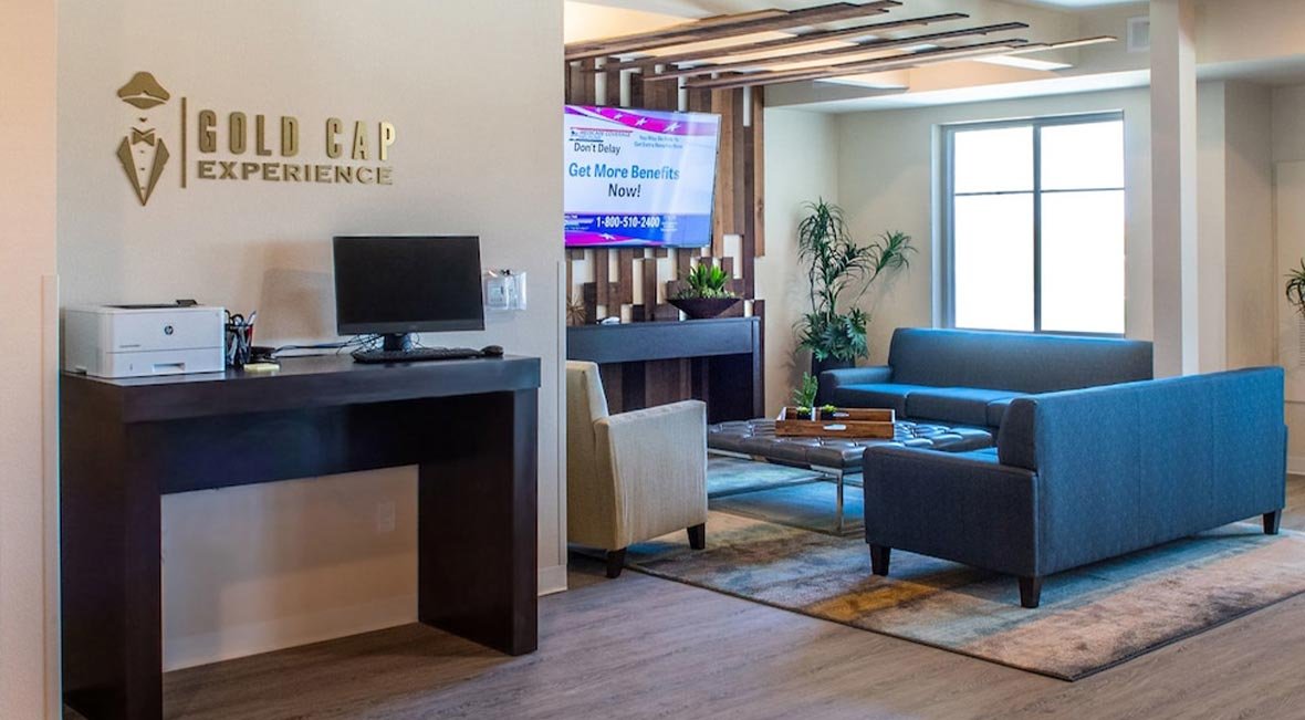 Reception area with a logo that says 'Gold Cap Experience', a monitor on a dark wood desk, and a sitting area with blue and beige chairs next to large windows.