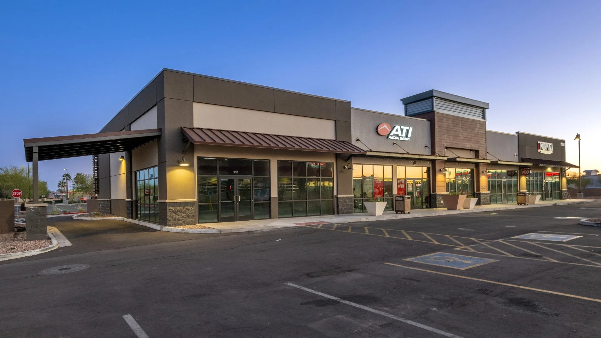 Empty parking lot in front of a strip mall at dusk, with stores including ATI Physical Therapy and Mod Pizza, illuminated by exterior lights.