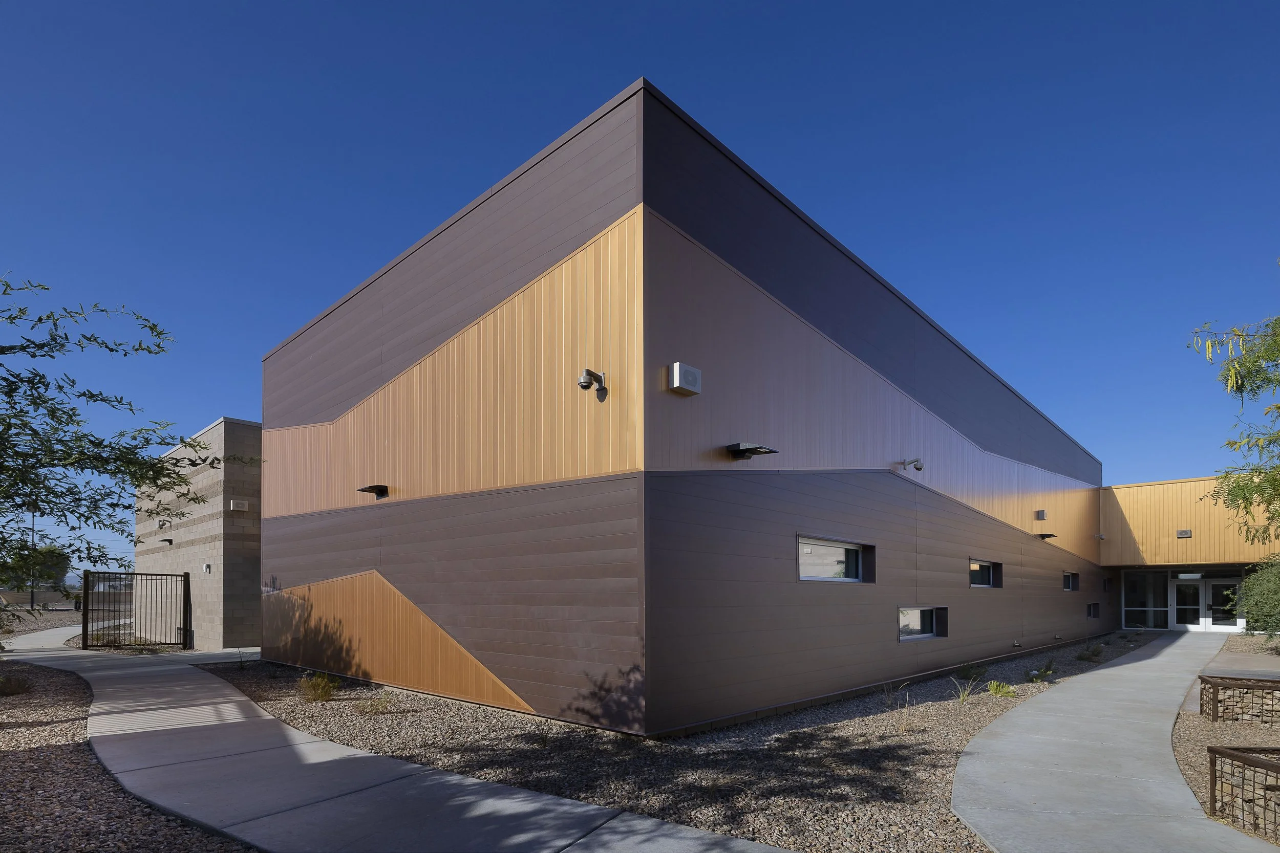 Modern multi-story building with brown and tan exterior walls, small windows, and a curved concrete walkway in front, under a clear blue sky.