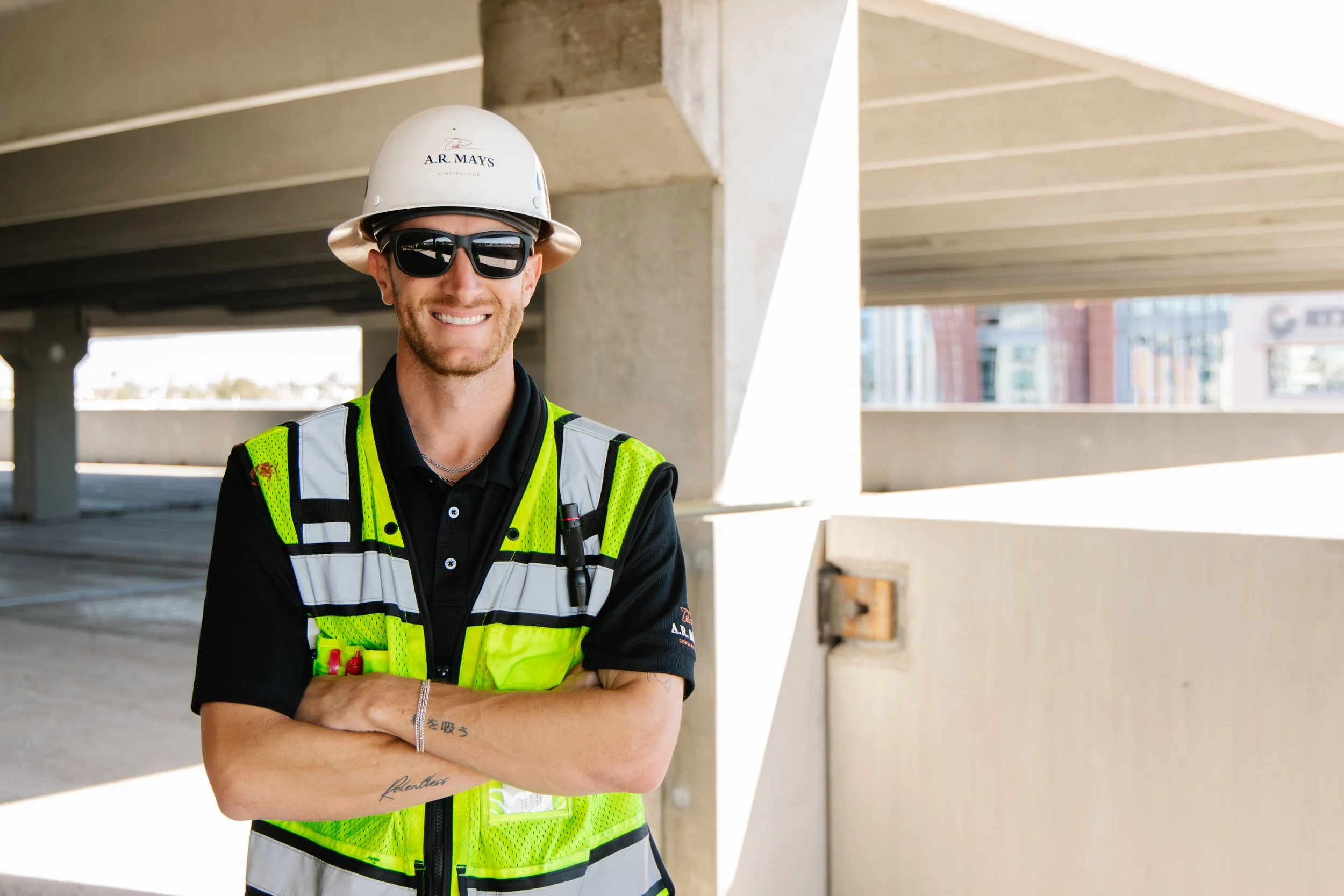 A construction worker wearing a white hard hat, black sunglasses, a black polo shirt, and a bright yellow safety vest with reflective strips, standing in an outdoor parking structure with arms crossed and smiling.