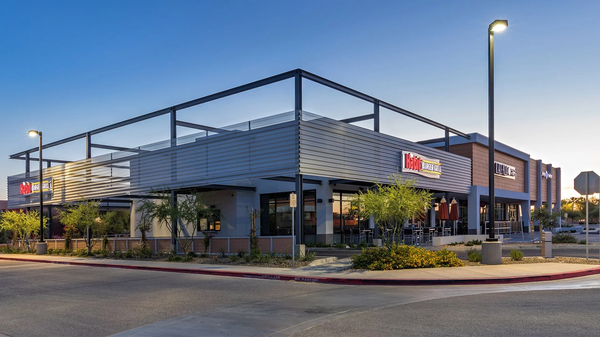 Exterior of a modern commercial building with signs for Habibi Burger Grill and other establishments, featuring outdoor seating, trees, and street lighting at dusk.