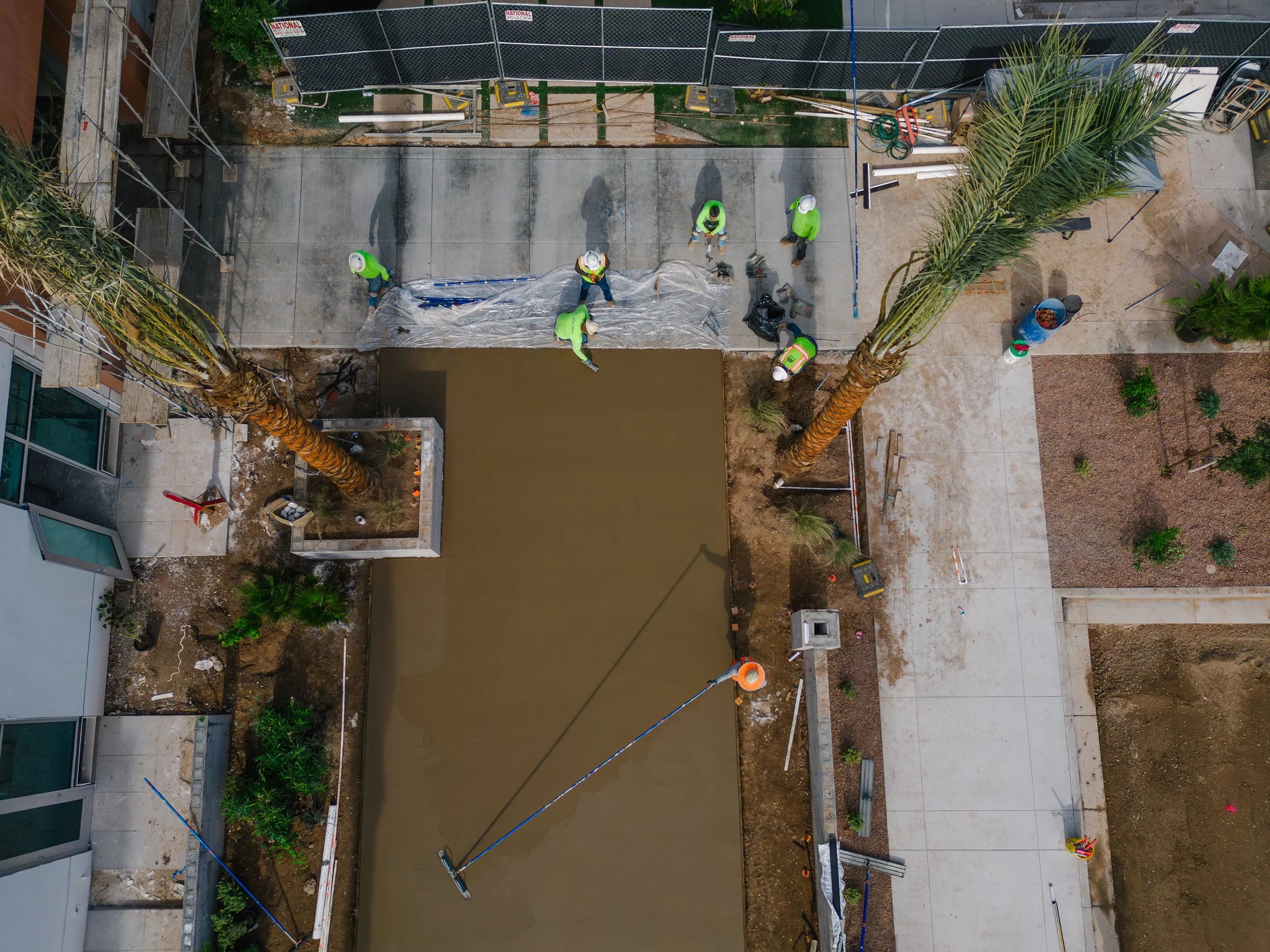 Construction workers are pouring and leveling concrete in an outdoor area, surrounded by construction equipment, tools, and partially finished surfaces.