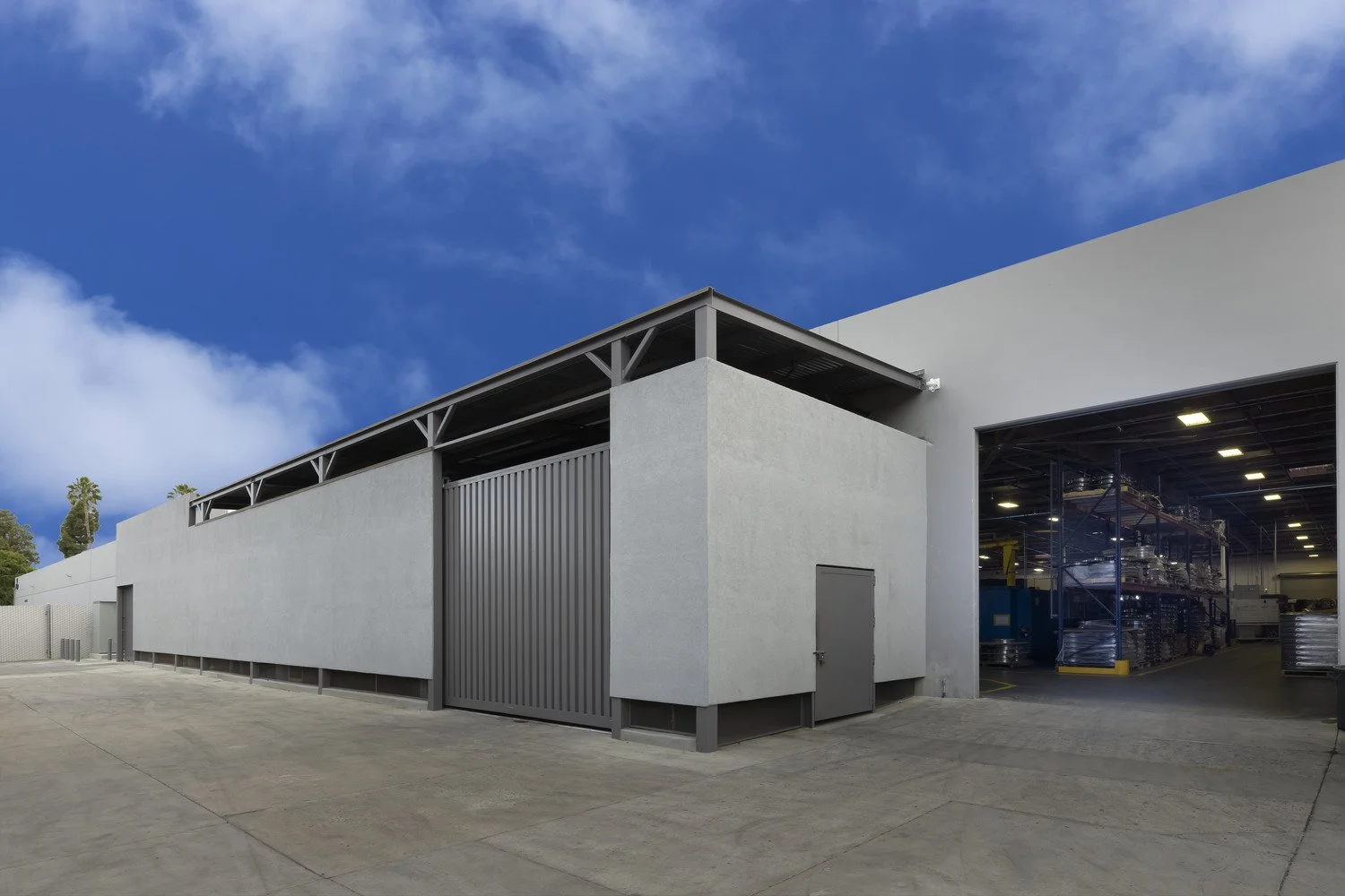 Exterior of a warehouse with a large sliding door open revealing shelves with supplies inside, against a bright blue sky with scattered clouds.