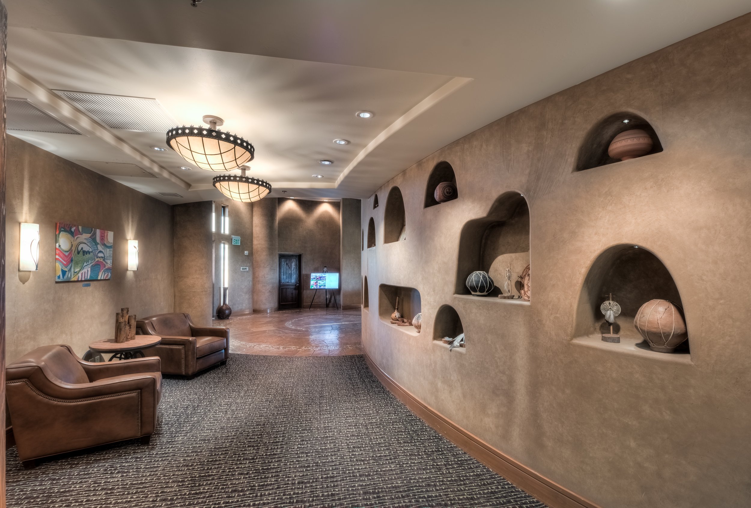 Lobby with two brown leather armchairs, wall-mounted art, wall sconces, built-in wall niches with decorative artifacts, wood door, elevator and a digital display.