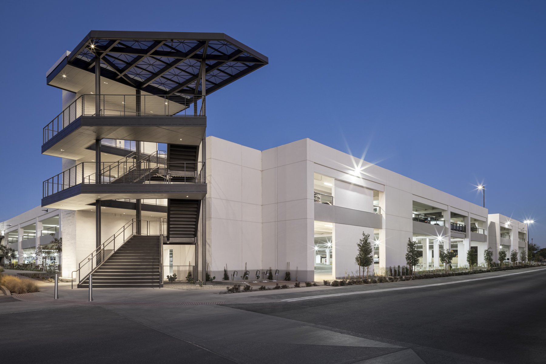 Modern multi-level parking garage with exterior stairs and a unique roof structure, illuminated at dusk, surrounded by landscaped trees and a paved road.