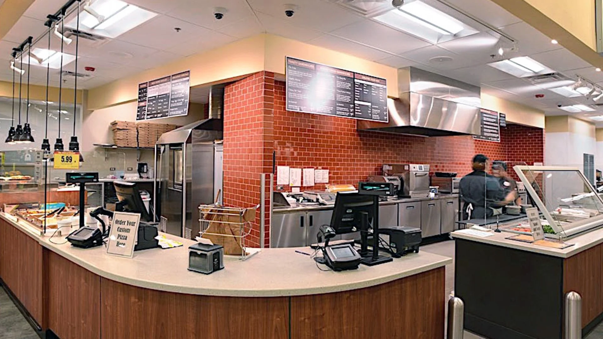 Interior of a pizza restaurant counter with a sign that reads 'Order Your Custom Pizza Here', stainless steel kitchen equipment, menu boards on the wall, and two staff members working behind the counter.