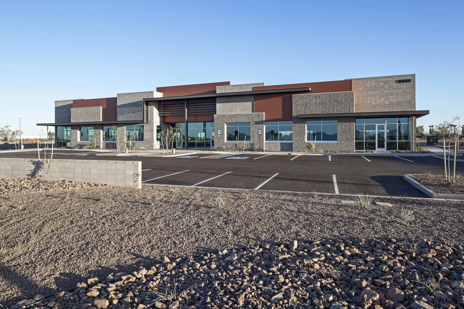 Modern commercial building with large glass windows and a multi-colored exterior, surrounded by a parking lot with marked spaces, in a desert-like landscape.