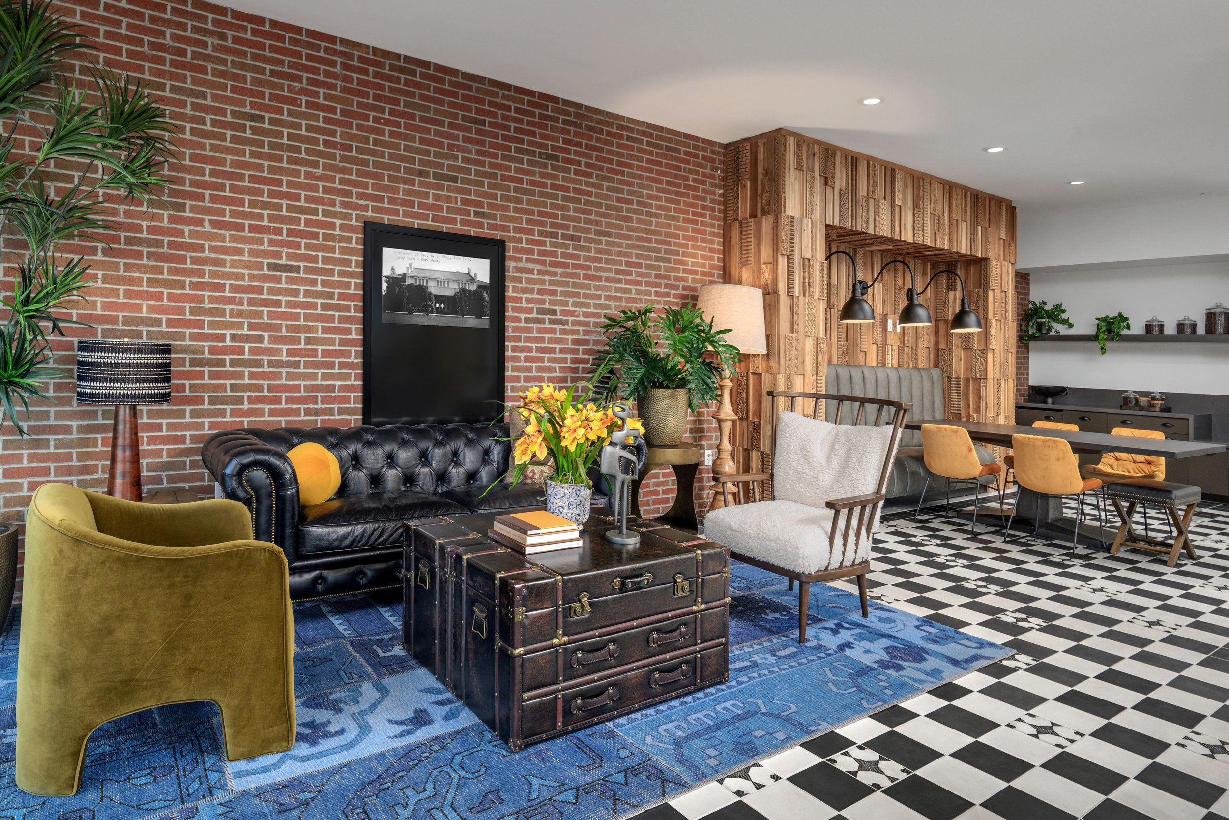 Living room with brick wall, vintage black leather tufted sofa, yellow armchair, wooden chair with pillow, coffee table with books and flowers, potted plants, floor lamp, and a dining area with yellow chairs, black table, and black-and-white checkere