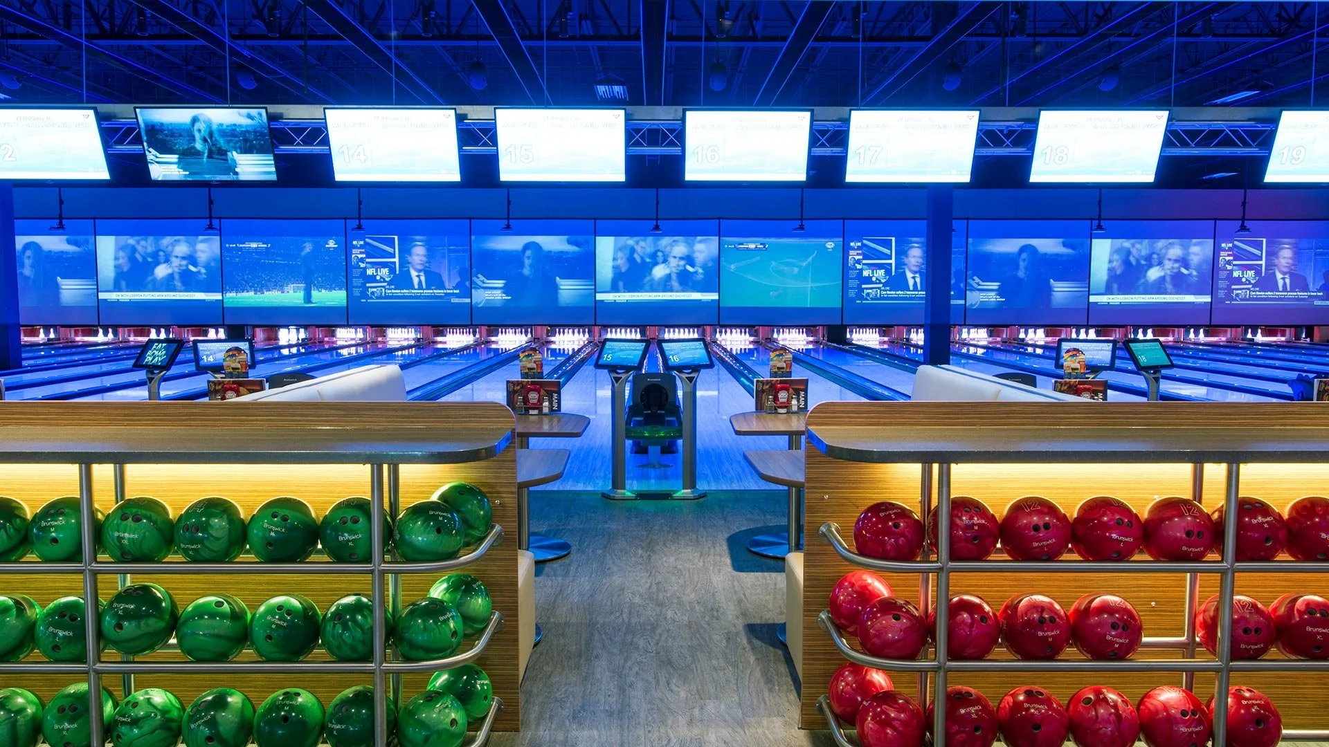 Inside a modern bowling alley with multiple lanes, digital score screens, and large TV screens displaying sports and news. Bowling balls in green and red are stored in racks in the foreground, and the lanes are illuminated with blue lights.