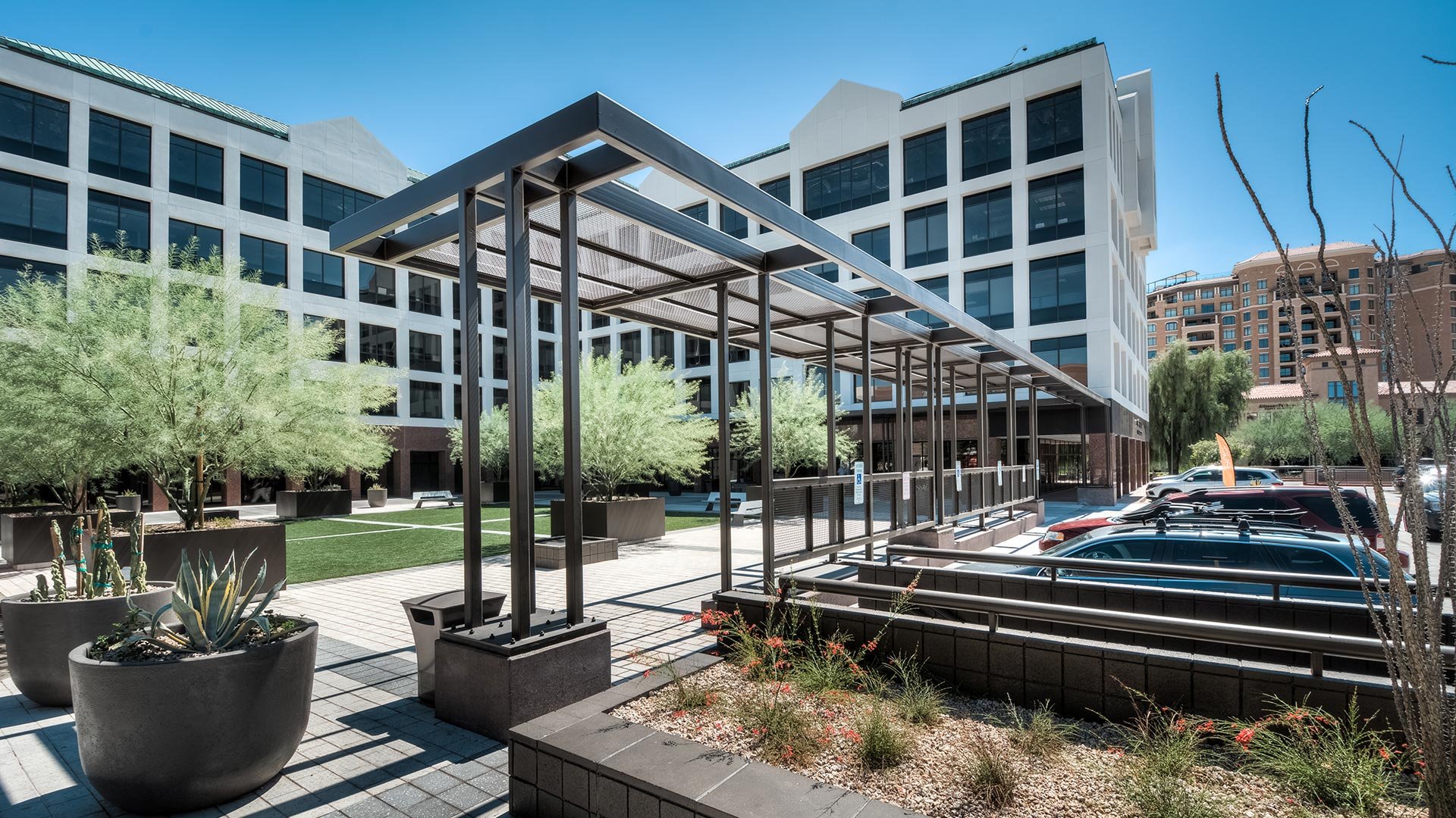 Modern urban courtyard with potted plants, trees, and a covered walkway, surrounded by multi-story office buildings and a parking lot filled with cars.