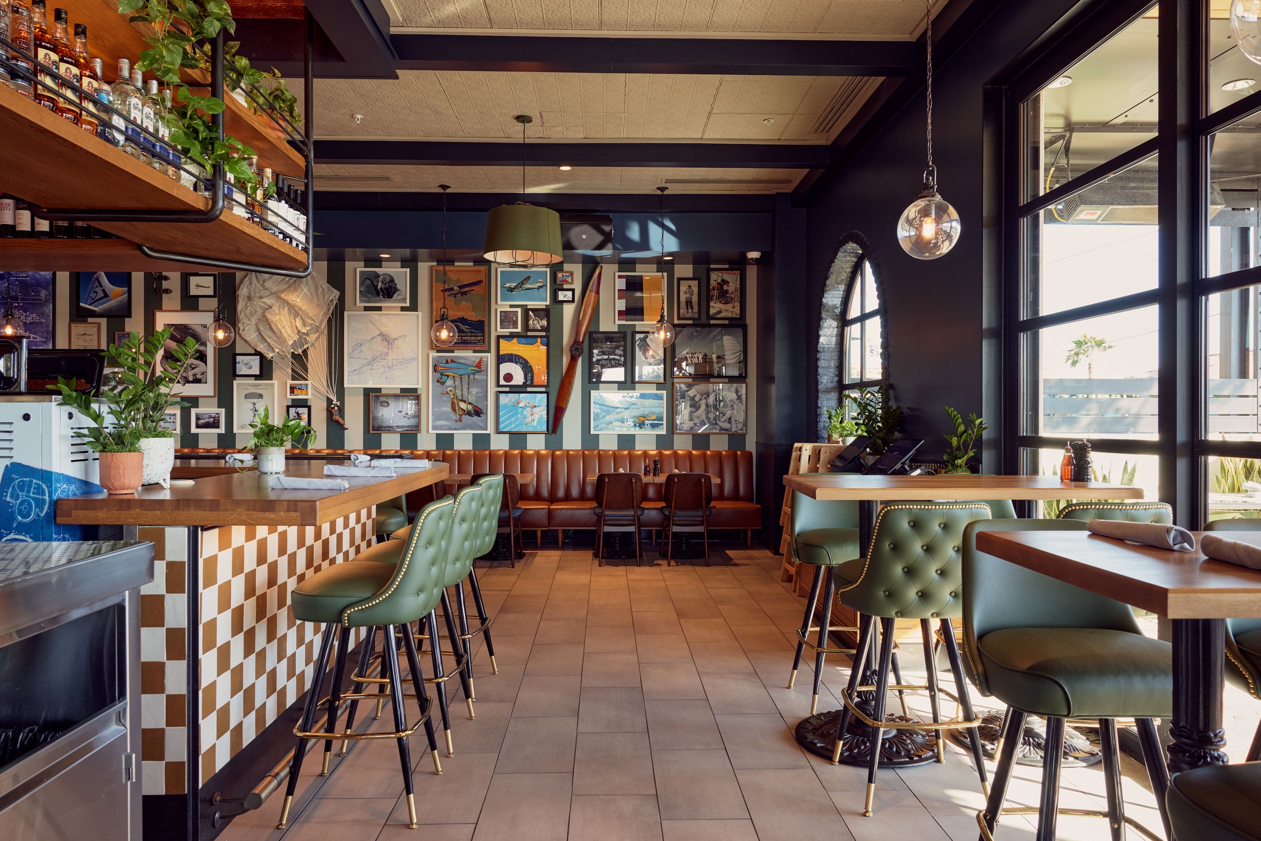 Interior of a cozy restaurant with a bar, green upholstered chairs, and wall decor including pictures and vintage aviation items, illuminated by natural light from large windows.