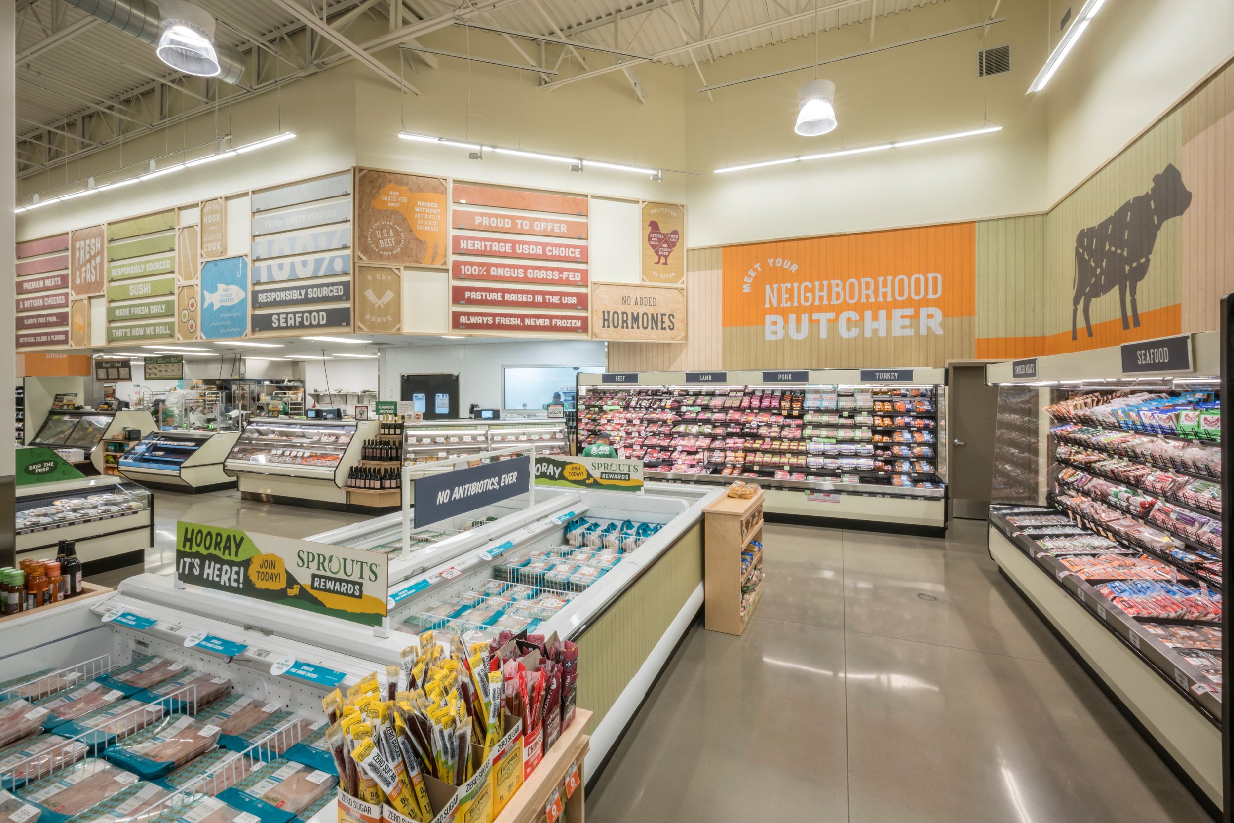 Interior of a grocery store with sections labeled 'seafood', 'pork', 'lamb', 'turkey'. Signs promoting local products and hormone-free meat. Fresh meat and seafood displays. Bright lighting, polished concrete floors, and colorful signage.