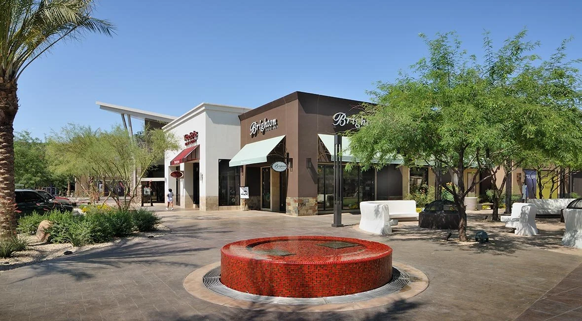 Outdoor shopping mall area with stores and seating, including a red tiled fountain, trees, and bench seating under clear skies.