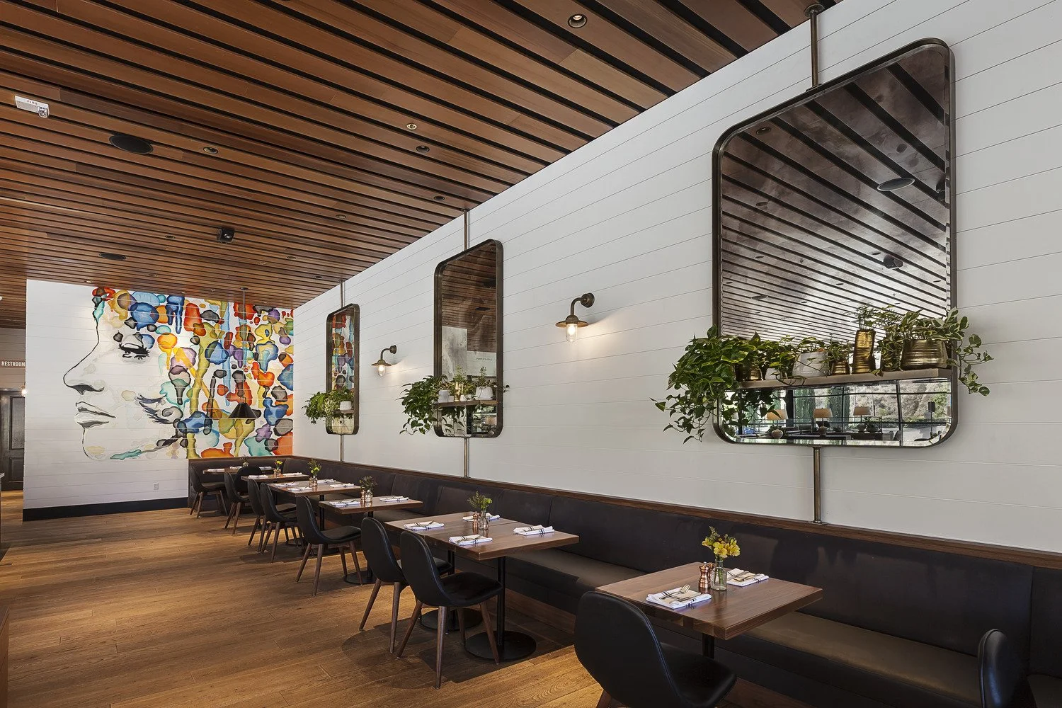 Interior of a modern restaurant with wooden floors and ceiling, white shiplap walls, black chairs, and tables set with napkins and small vases of flowers. There is a colorful abstract mural of a woman's face on the wall.
