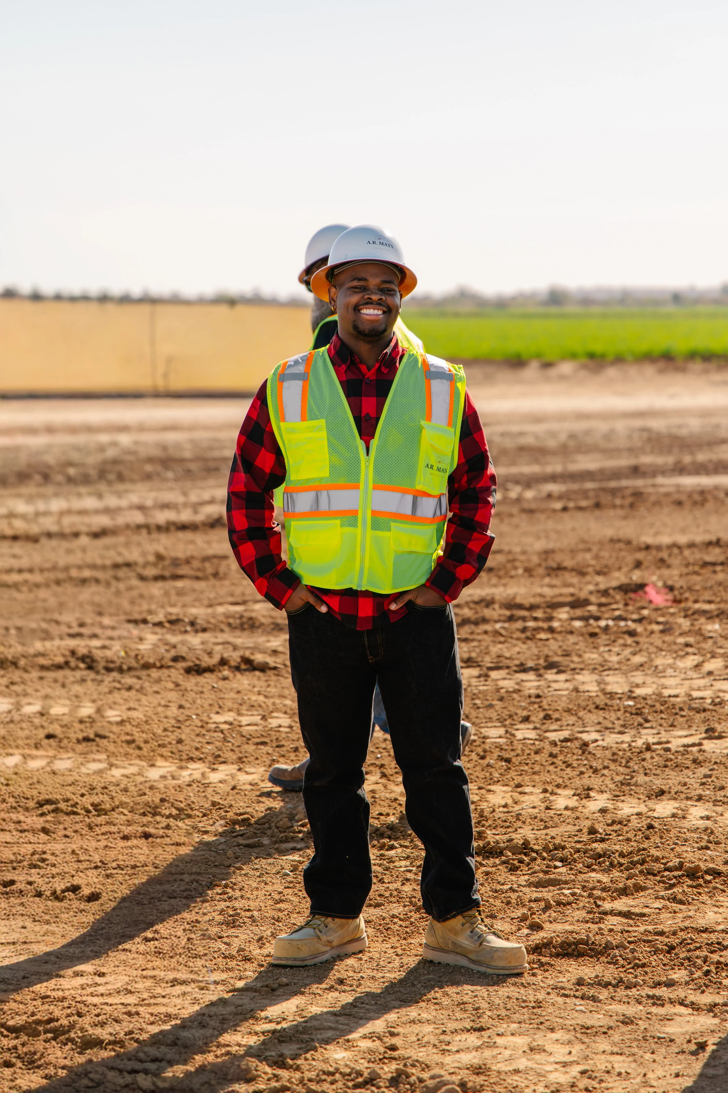 A smiling man wearing a yellow safety vest and a white construction helmet standing on a construction site with dirt ground and green fields in the background.