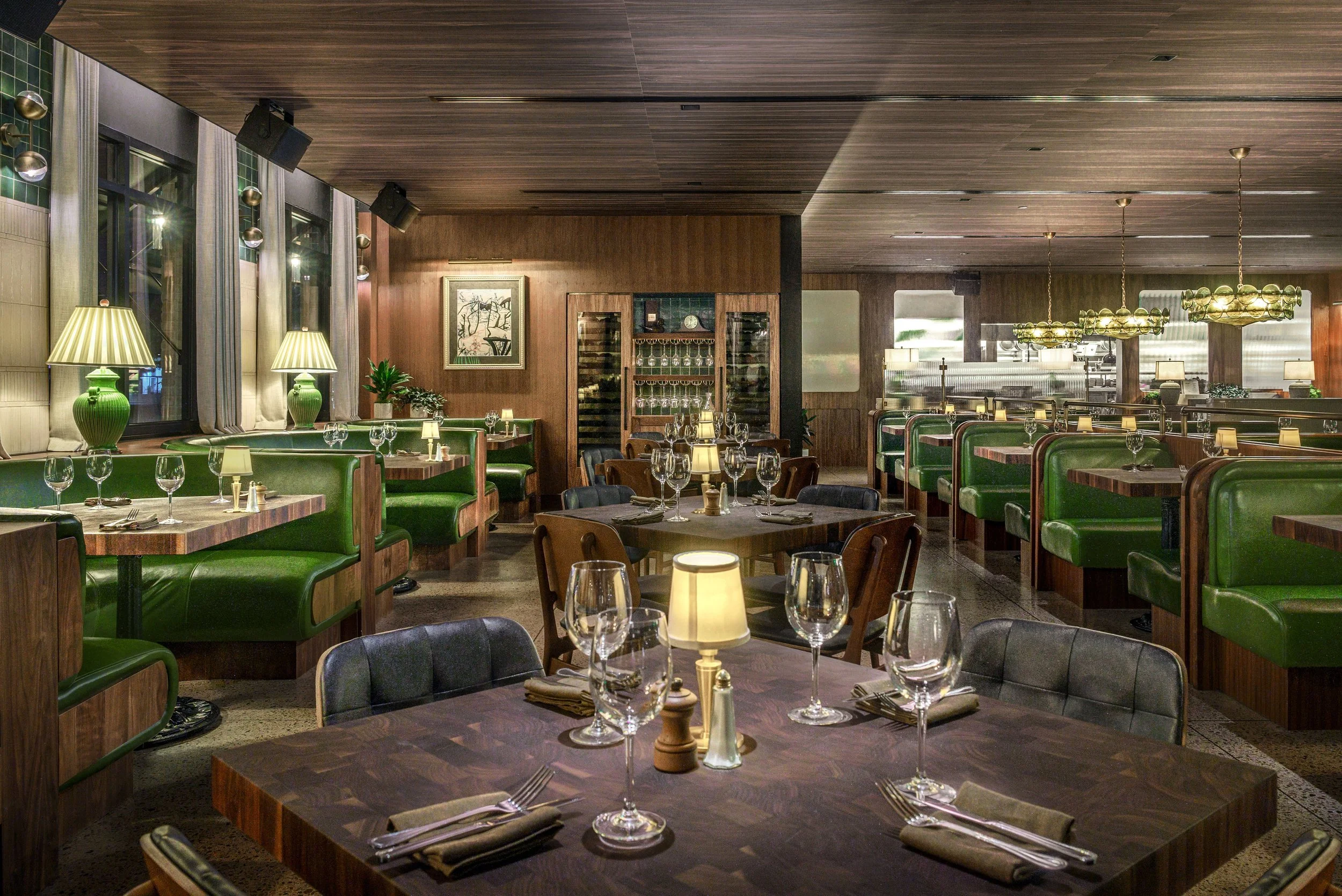 Interior of a restaurant with green booth seating, round tables with wine glasses, tables with lamps, and wooden walls with artwork and a wine cabinet.