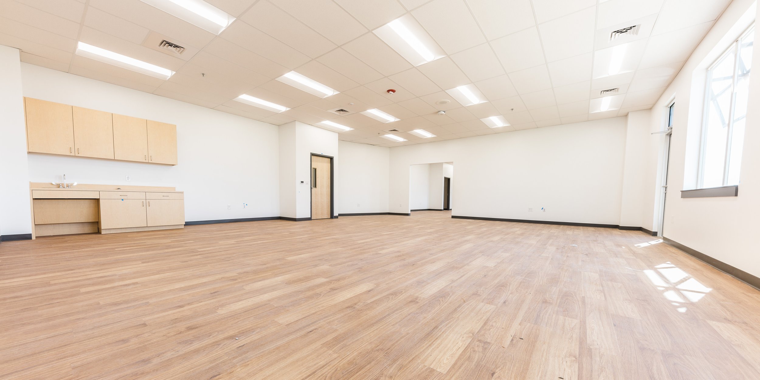 Empty room with wood flooring, white walls, large windows, and ceiling lights, with a small kitchenette on the left side.