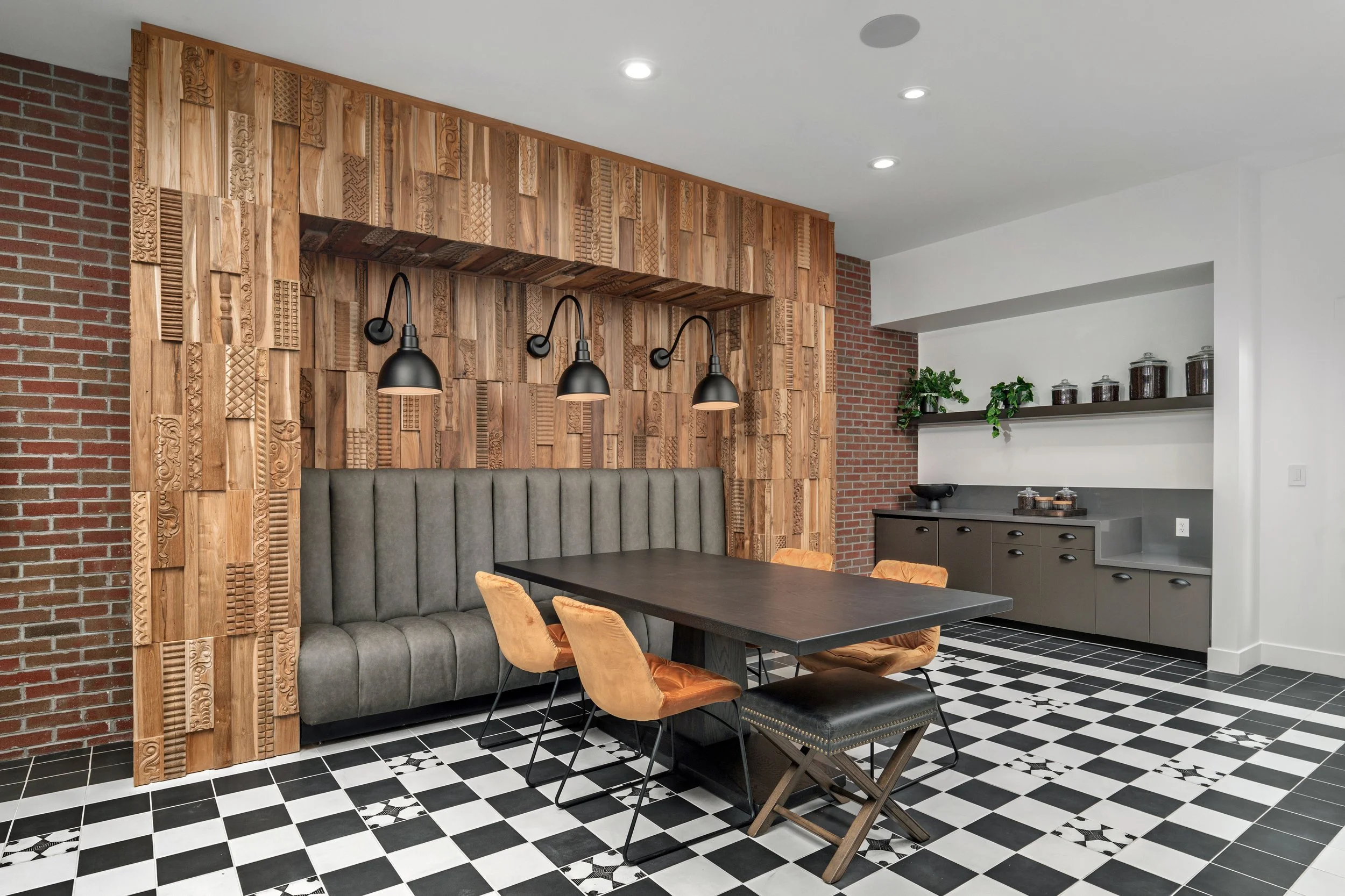 Modern dining area with a black table, tan and black chairs, gray upholstered bench, wooden accent wall, black wall lamps, and a sideboard with storage and decorative jars, black and white tile floor.