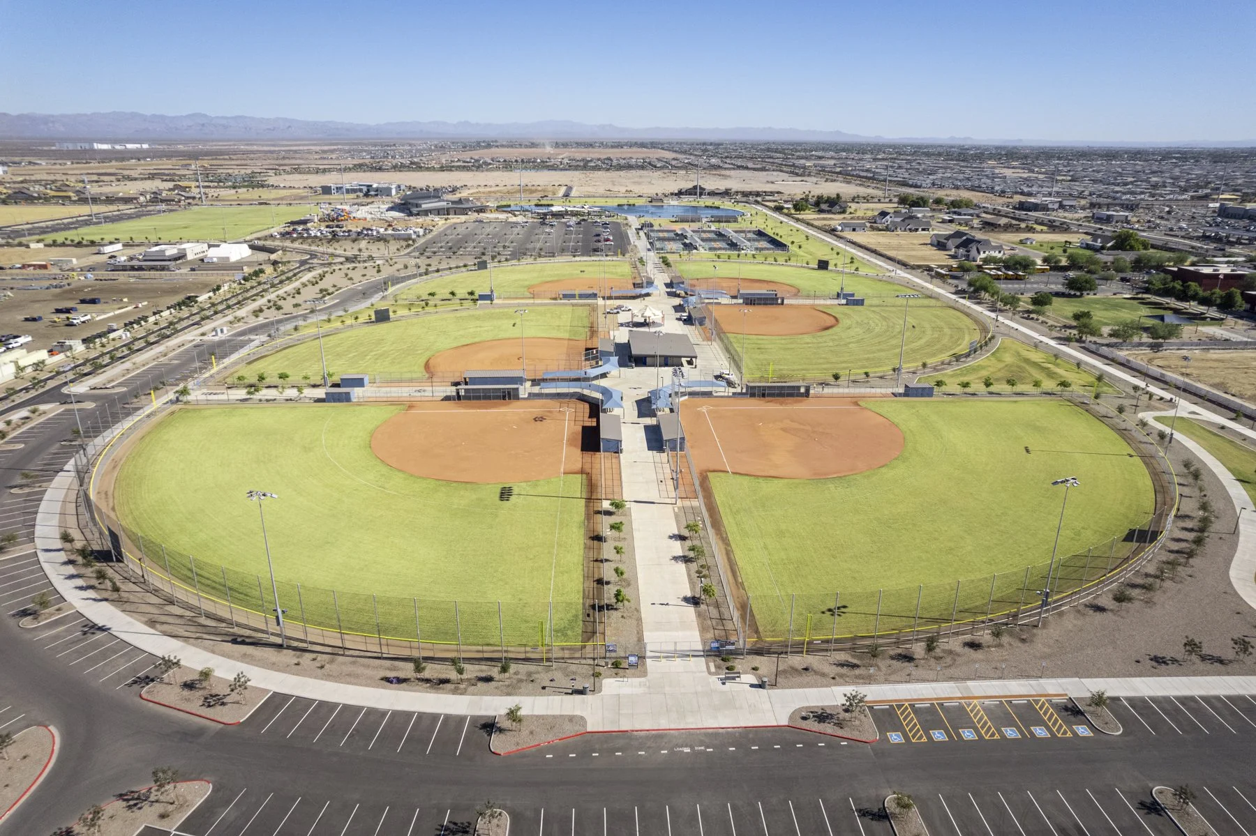 Aerial view of a baseball complex with four fields, parking lots, and surrounding roads on a clear day.