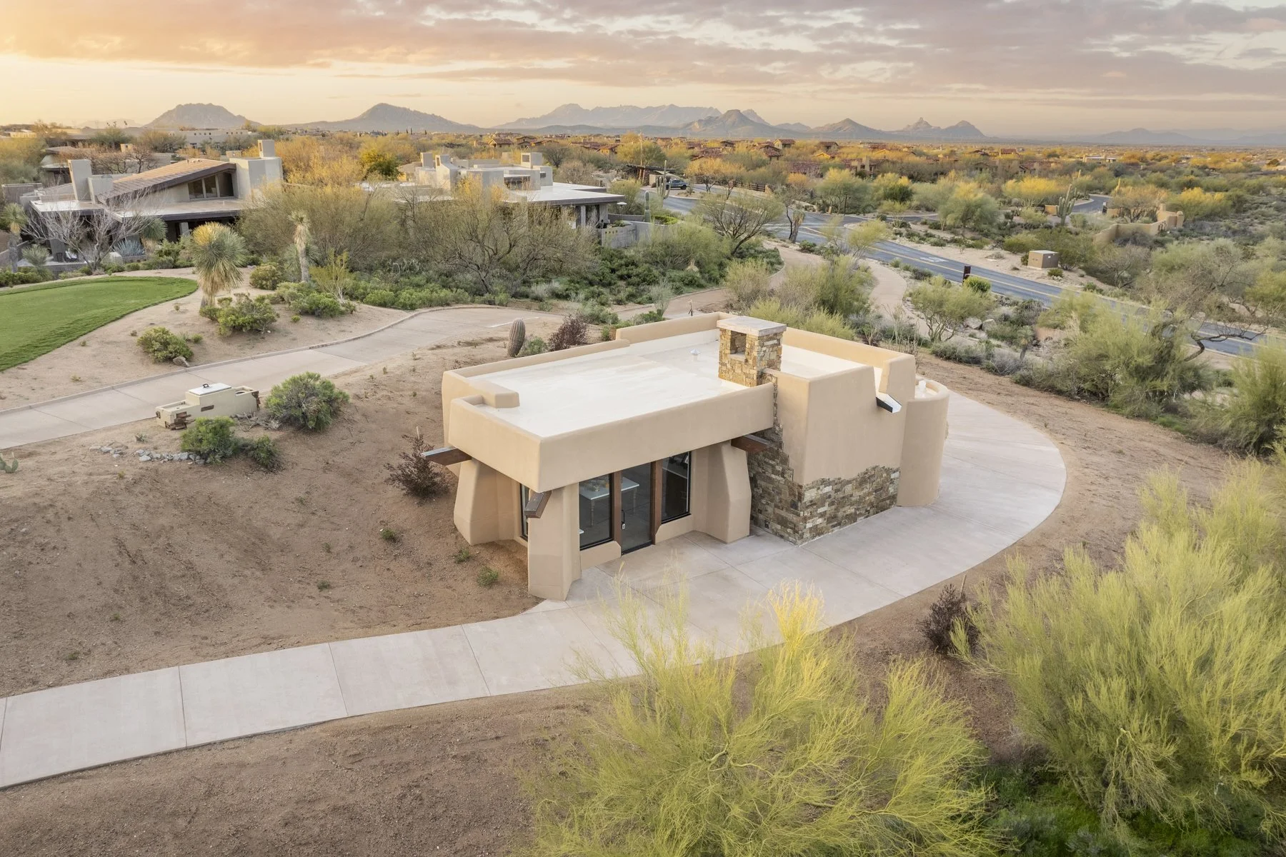 A modern house in a desert landscape with mountain views, a curved driveway, and sparse vegetation including bushes and cacti, during sunset.