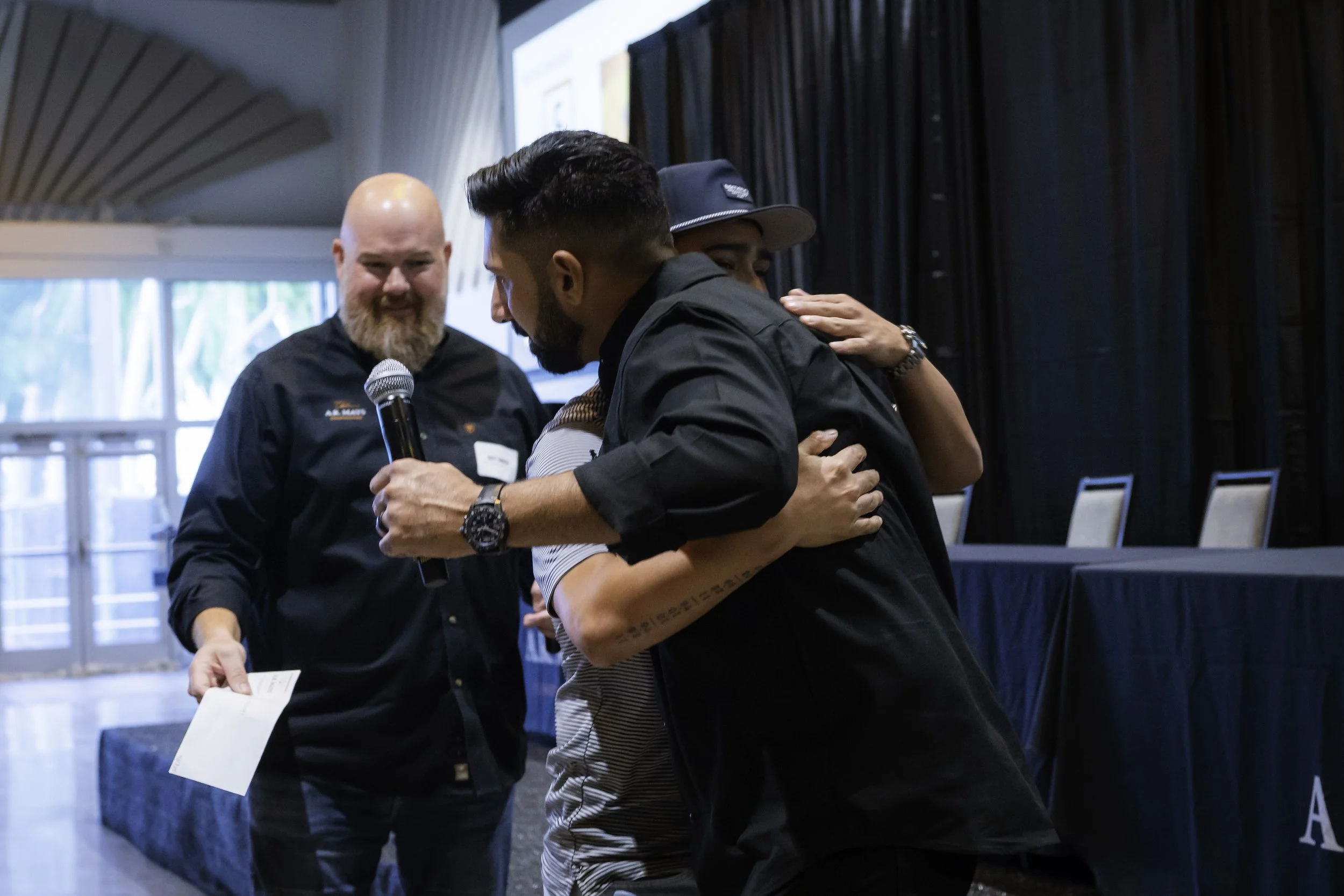Three men engaged in a warm embrace at an indoor event, with a background featuring dark curtains and large windows.