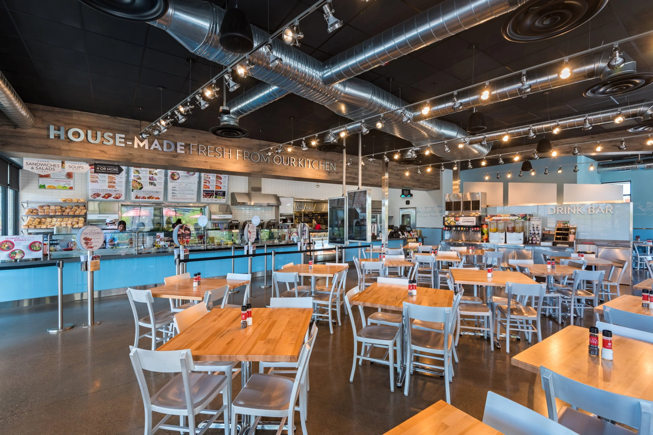 Interior of a modern casual restaurant with wooden tables and white chairs, showing a counter with food options and a drink bar in the background.