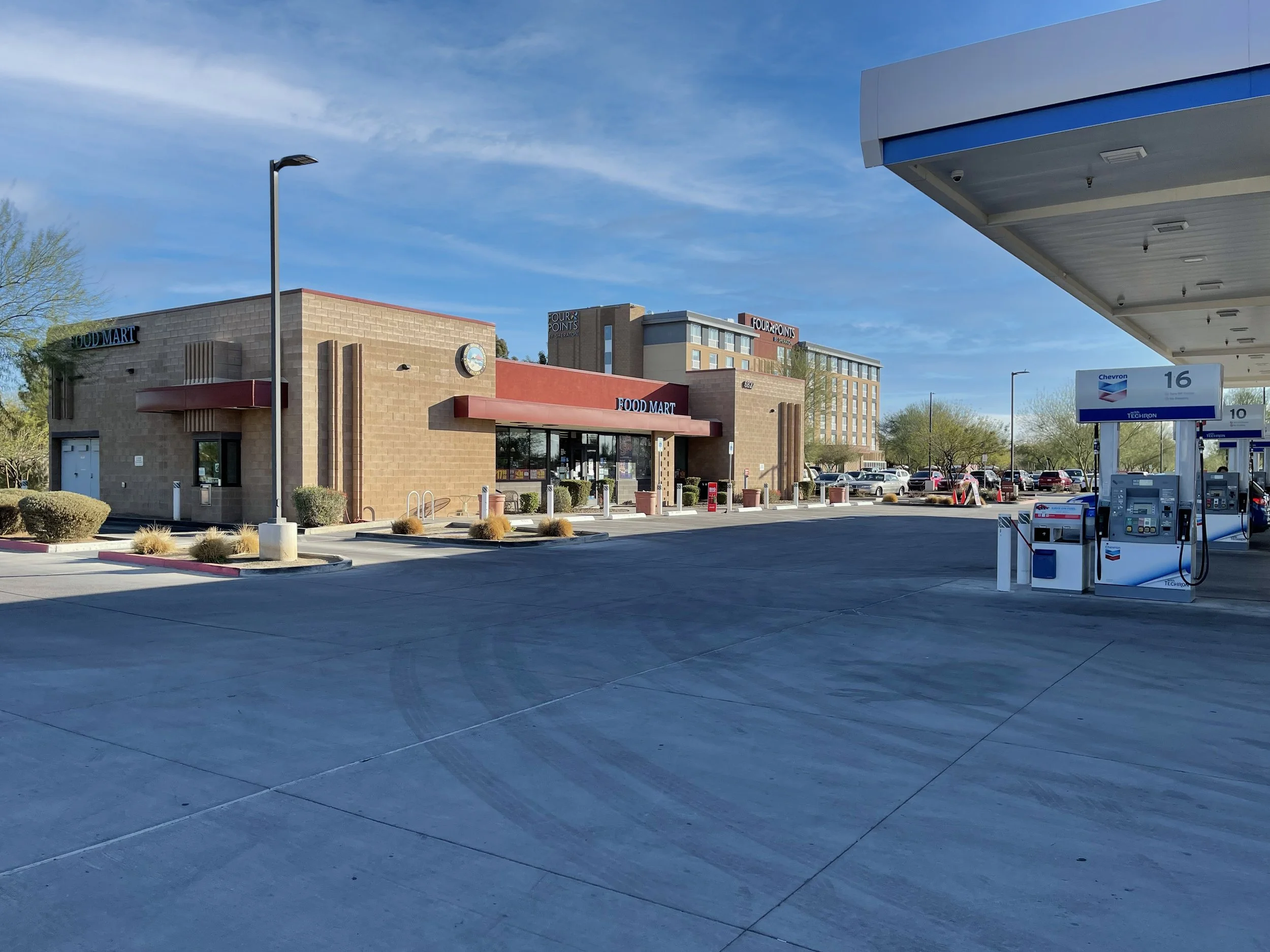Empty gas station with pumps under a canopy, a food mart store in the background, parking lot with cars, and a clear blue sky.