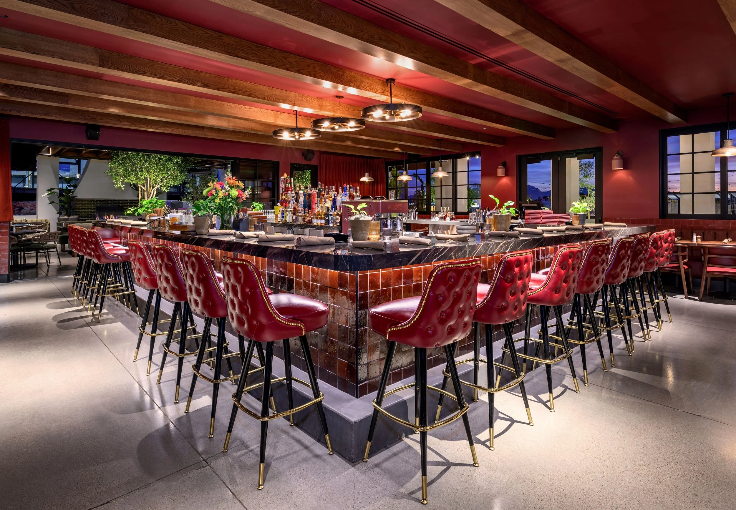 A stylish indoor bar with a large square marble-top counter, surrounded by red leather barstools with gold accents. The bar is well-appointed with various bottles of liquor, plants, and flowers. Large windows offer a scenic view outside, and modern p